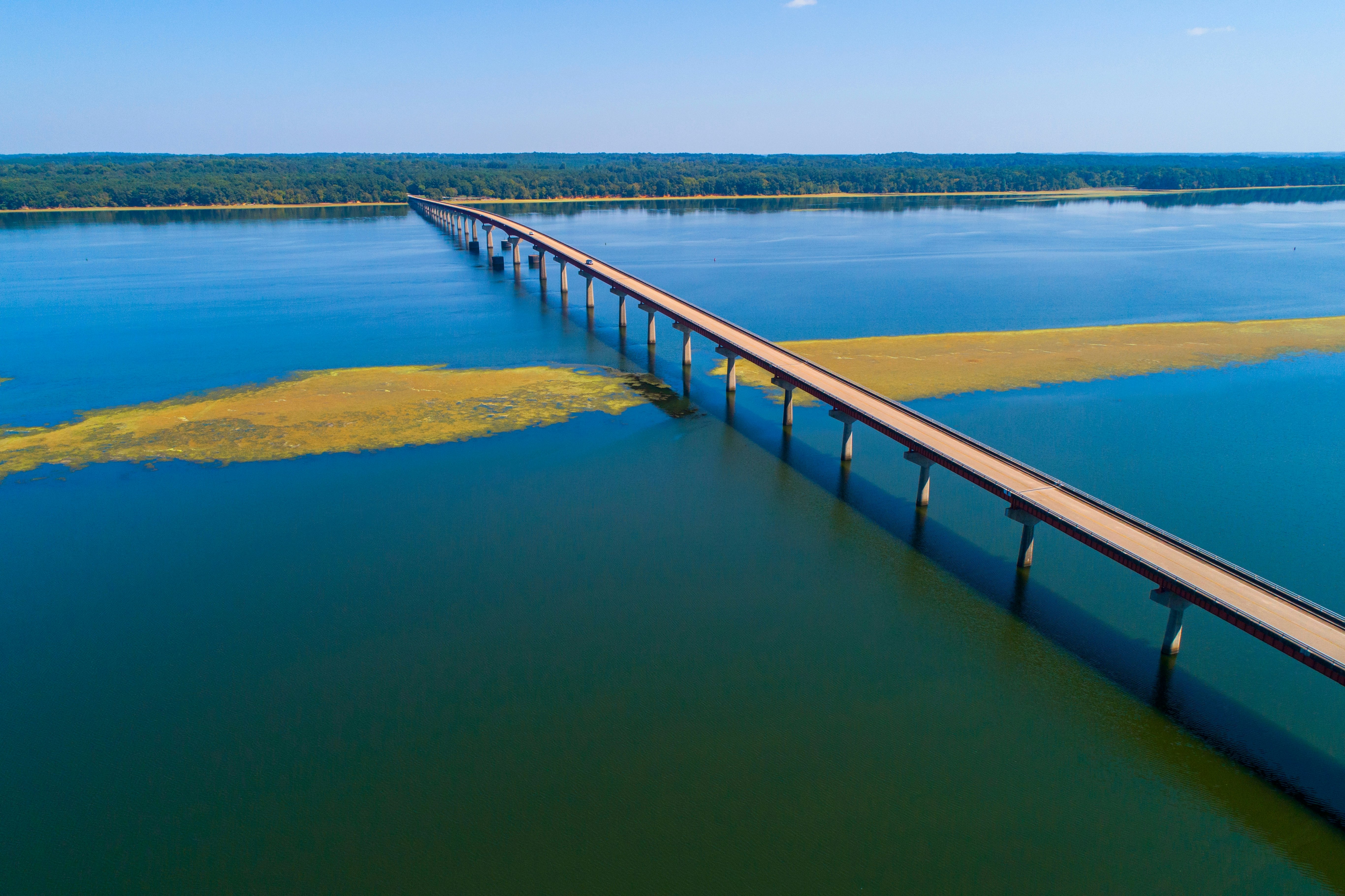 An aerial view of a long bridge spanning a wide river. Small islands are seen in the river, while trees line the riverbank in the distance.