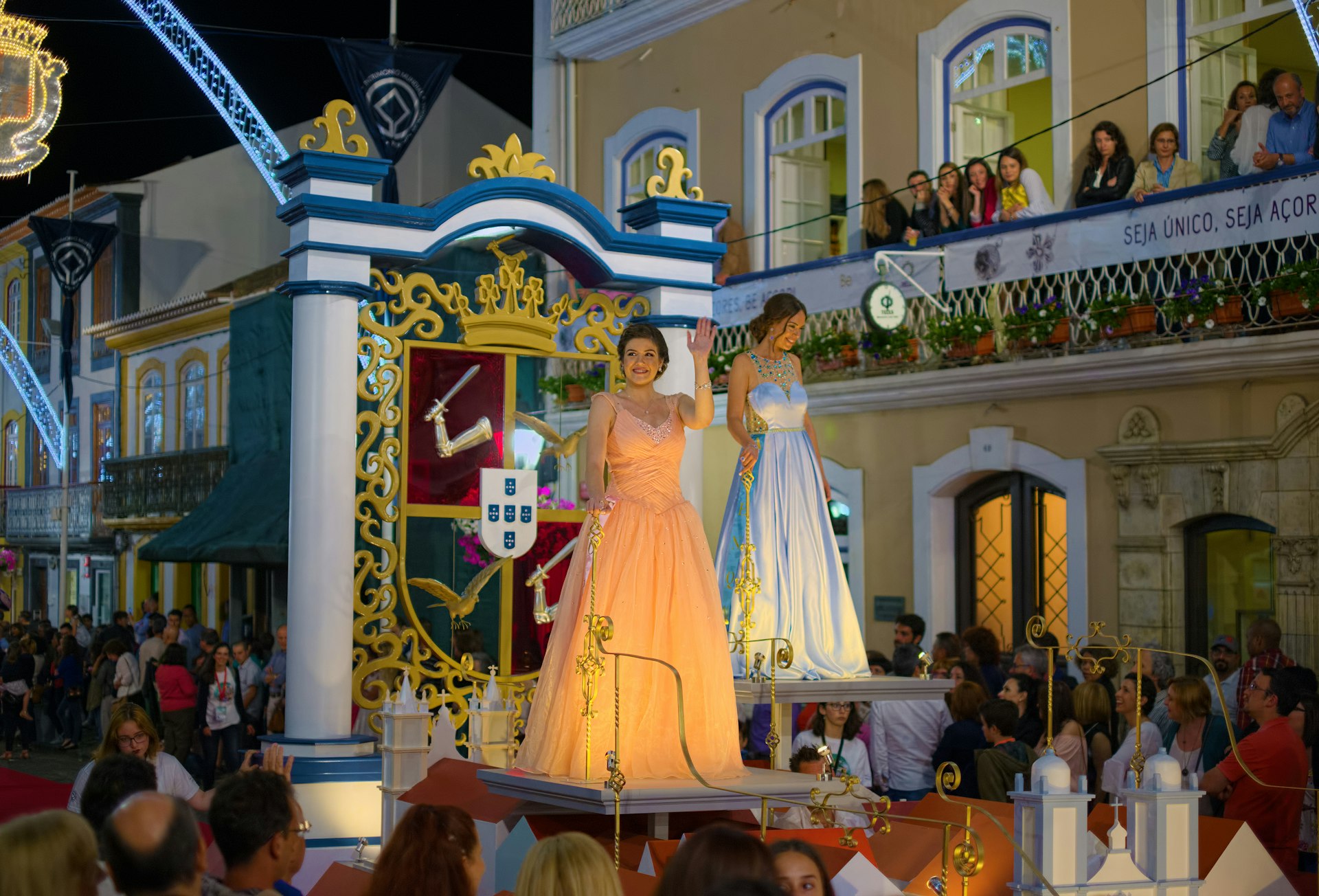 Crowds are watching as two women dressed in period gowns stand on a float during a parade through the streets of Angra do Heroismo, Terceira, for the Sanjoaninas festivities.