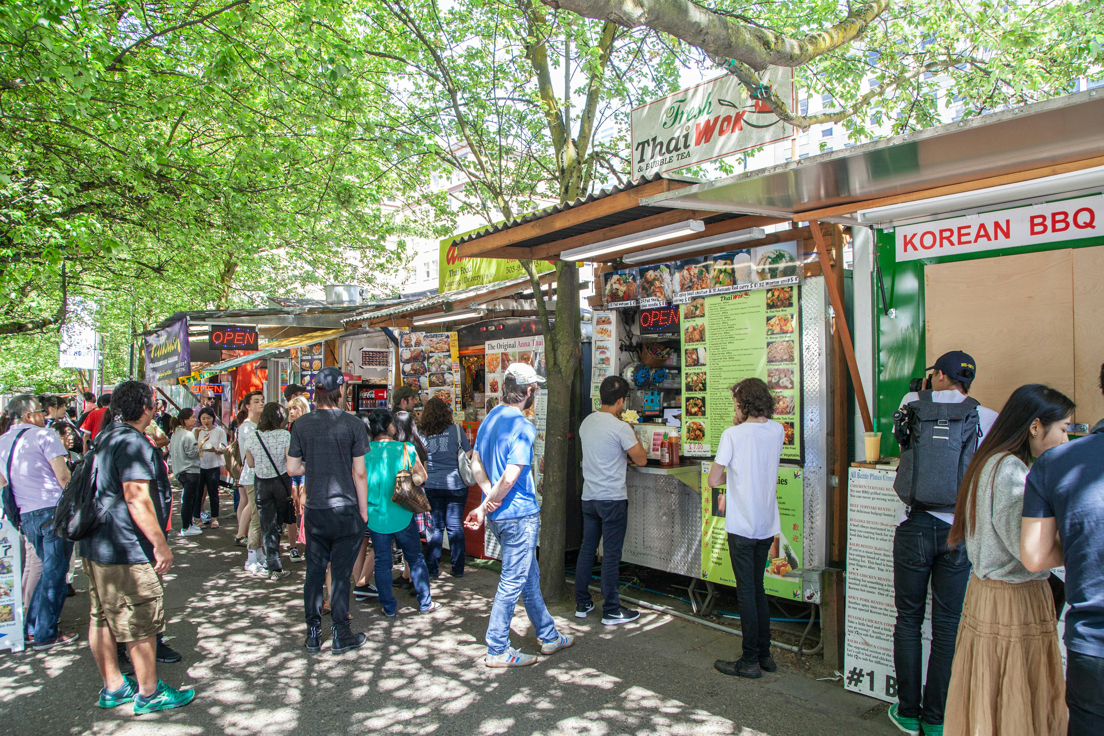 People line up to order Thai, Korean and other kinds of cuisine from a row of food trucks in Downtown Portland, Oregon. It is a sunny, warm day, with the people sheltered under branches with green leaves.