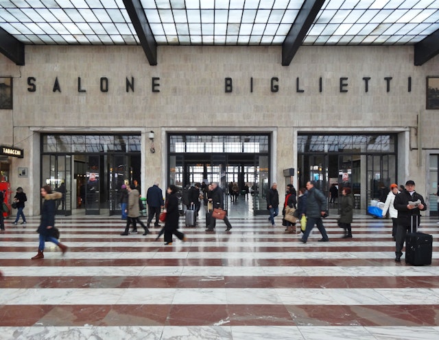 People carrying luggage blur past a station ticket office