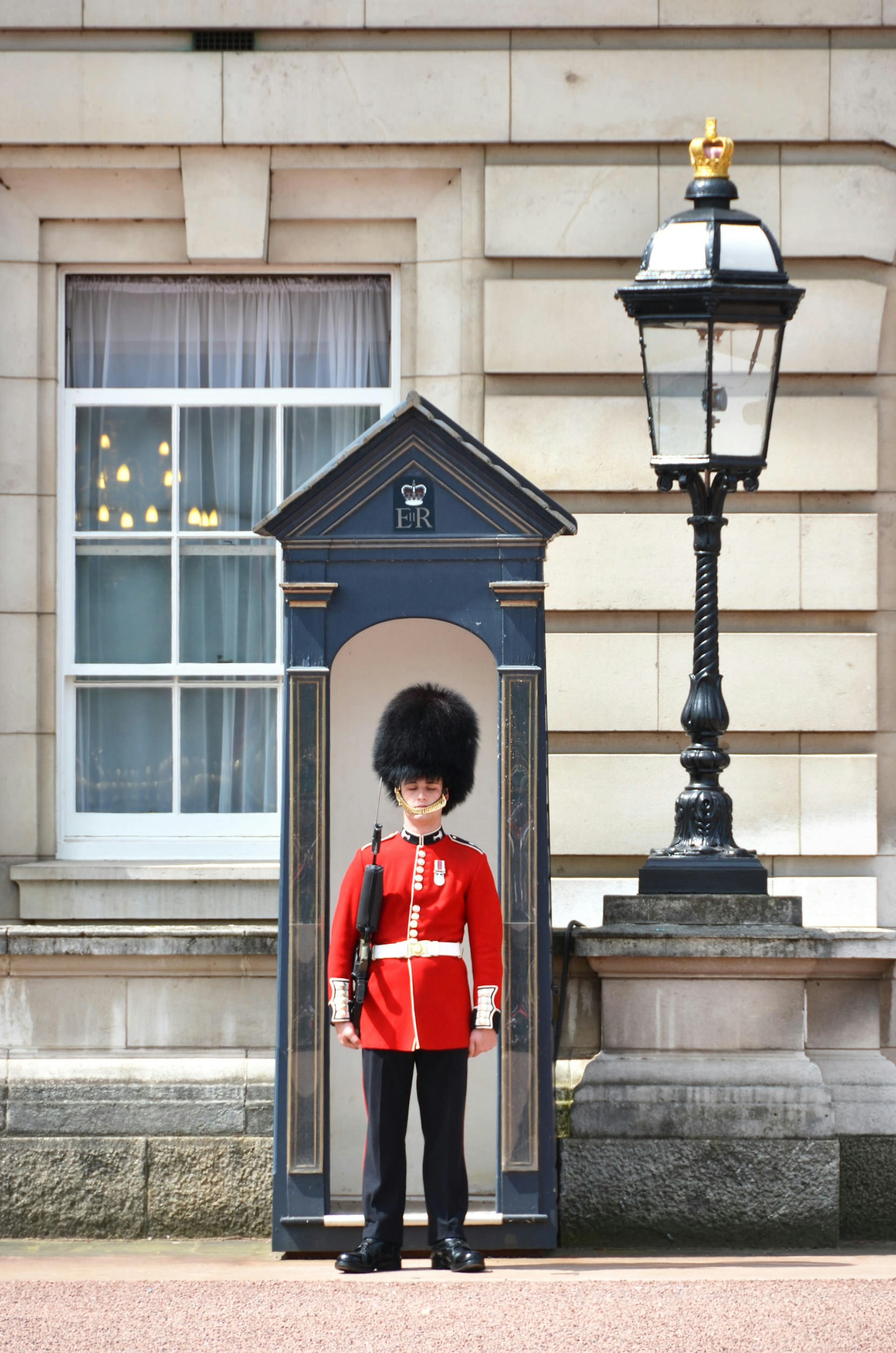 A guard in a fuzzy hat and a red jacket stands at attention in front of a booth and next to an ornate lamp