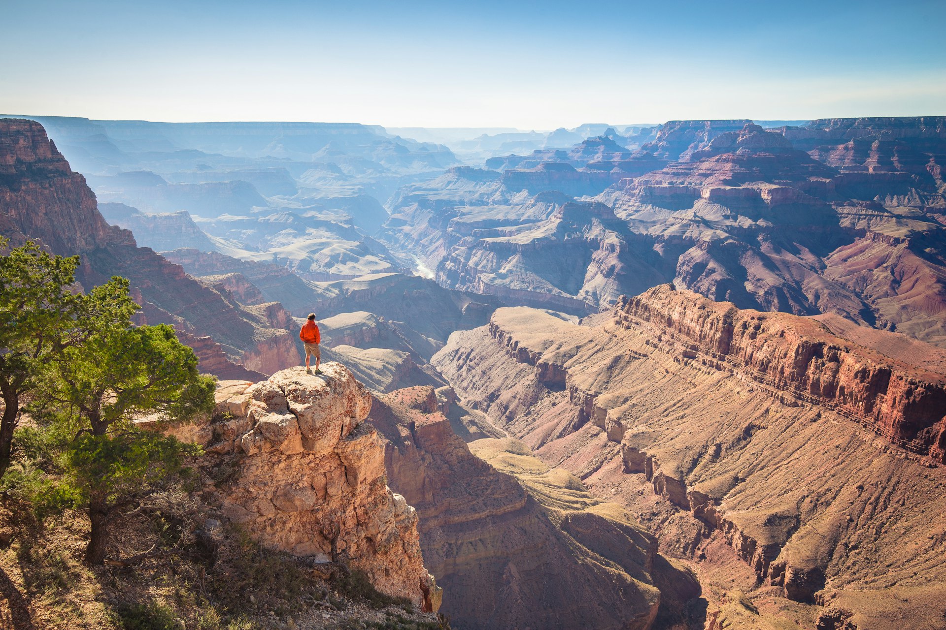 A solo figure stands on a rock looking out across a vast canyon as the sun shines down