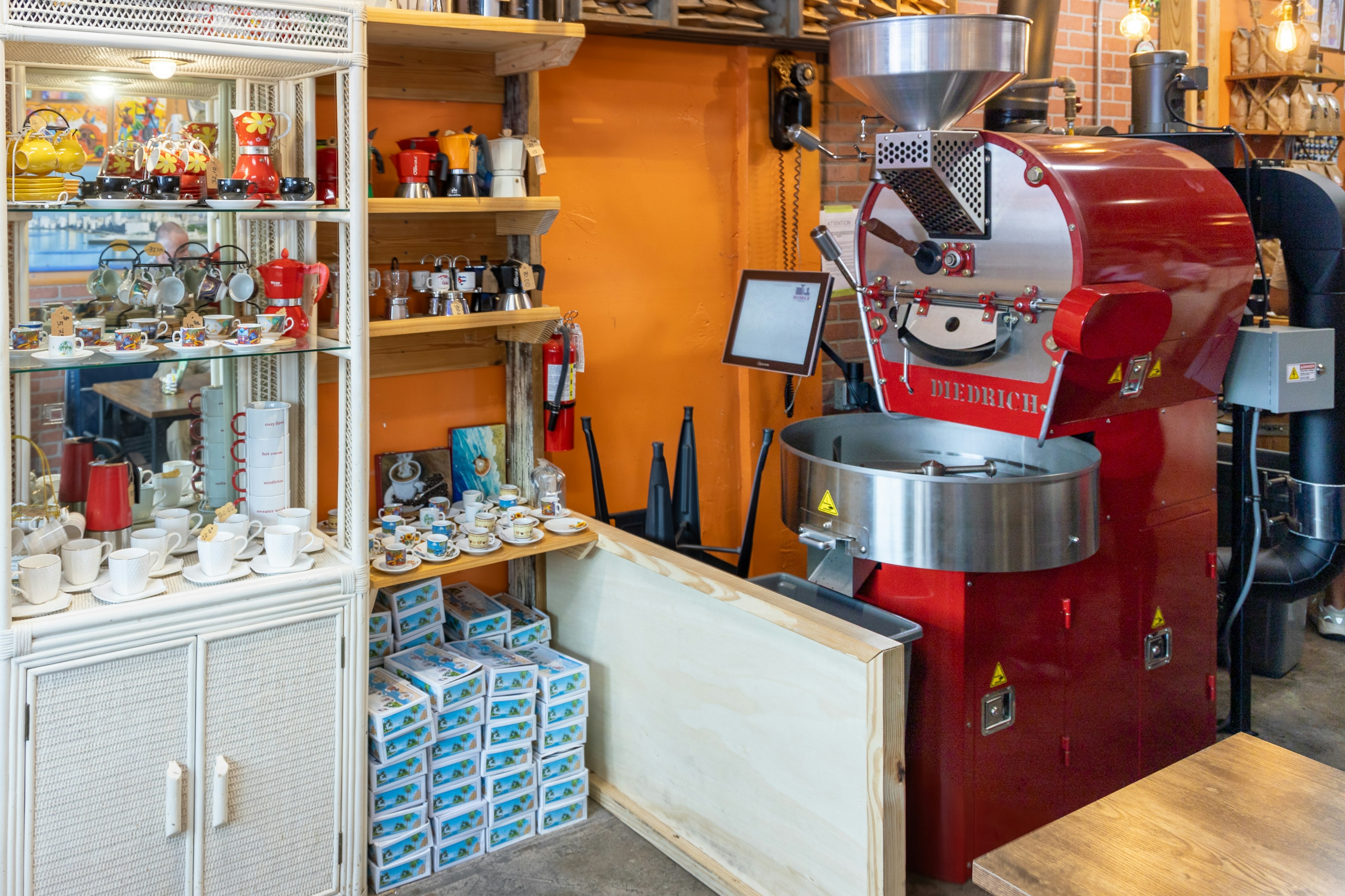 A display cabinet next to a large red coffee grinder is filled with espresso cups and coffee-themed ornaments for sale