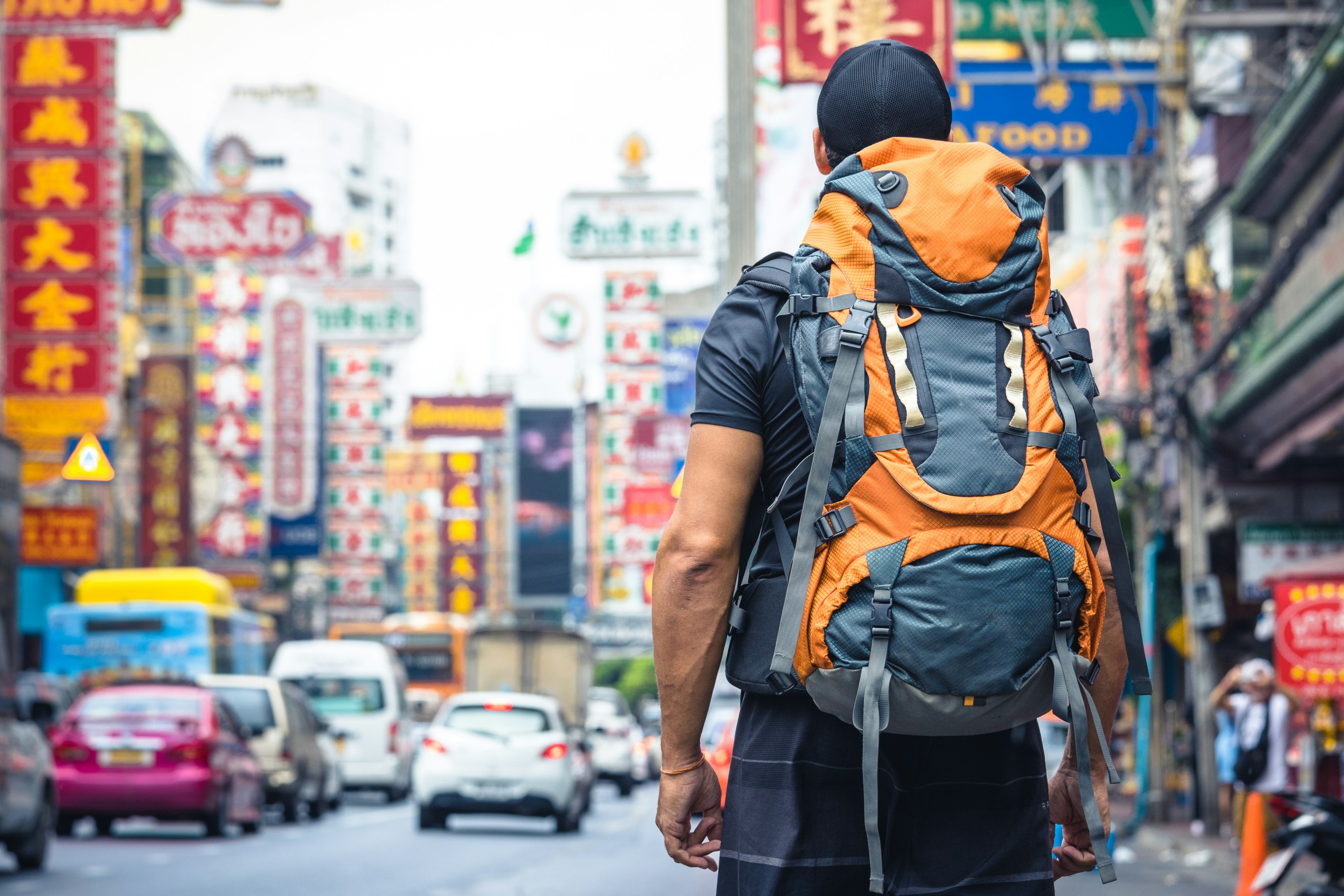 A male traveler wearing a large yellow backpack is standing on a very busy road in Bangkok, looking towards the traffic and many neon signs.