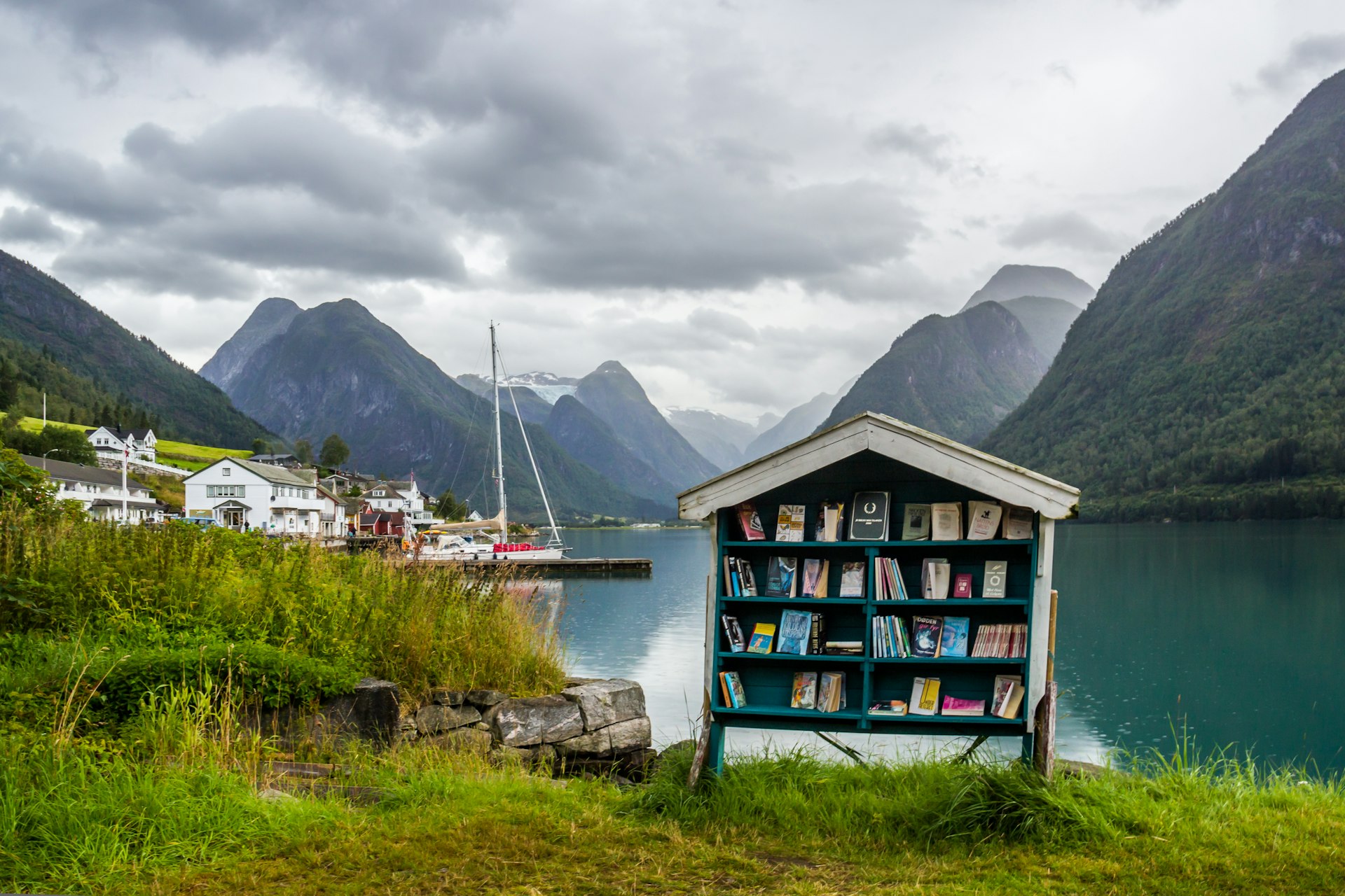 A cabinet full of books sits on the edge of a mountain-ringed fjord. 