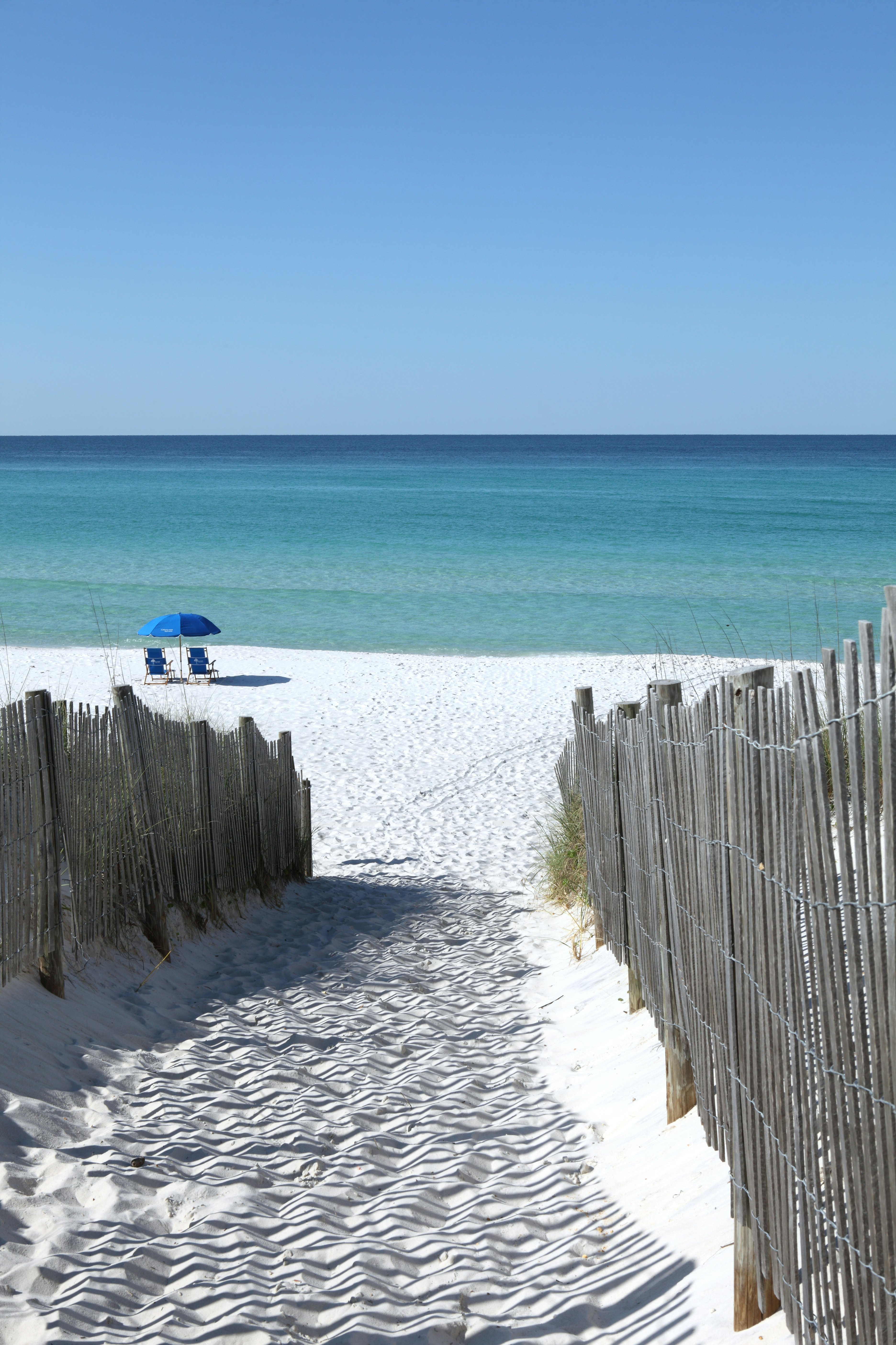 A sandy pathway leads down to a beach with a blue umbrella