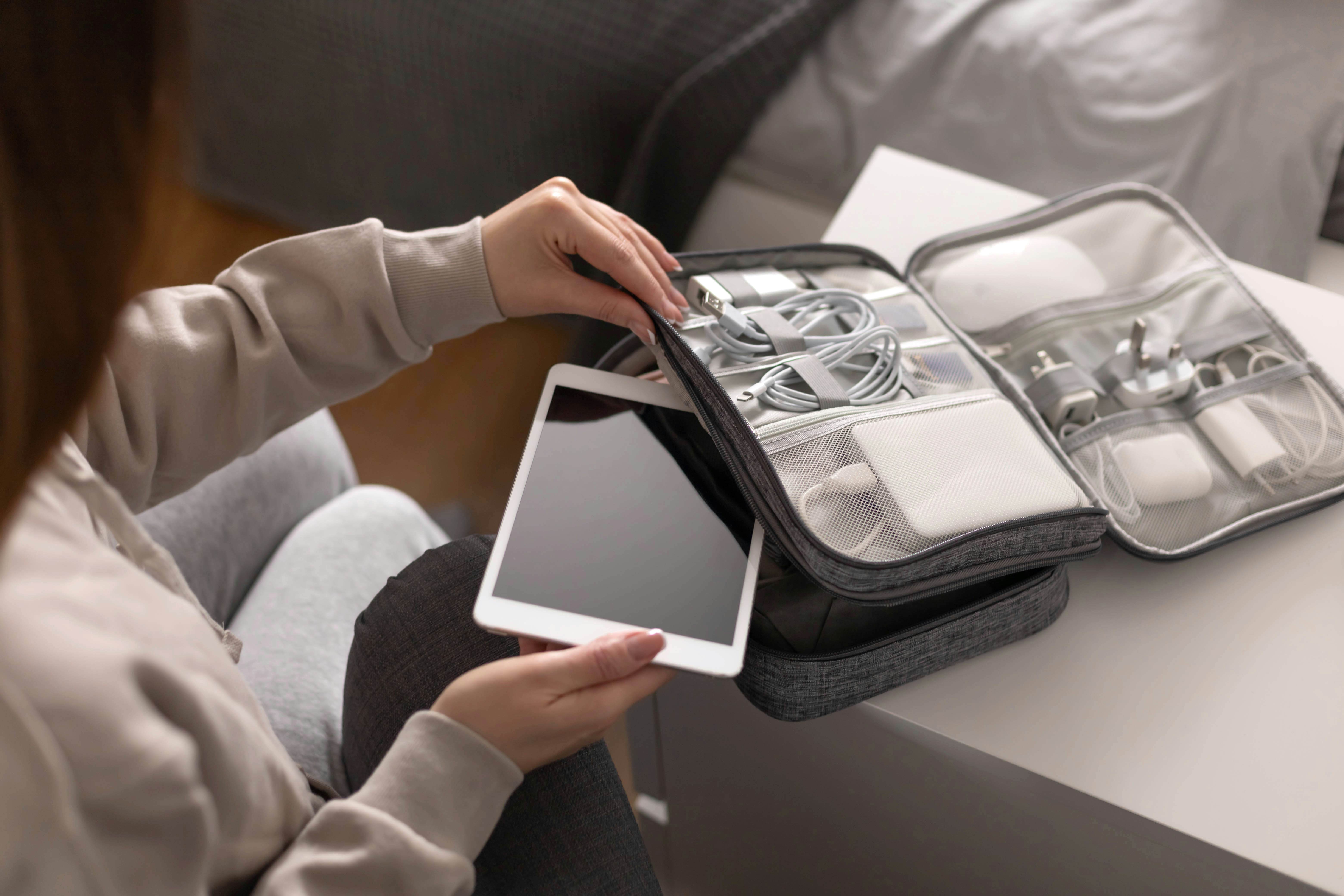 A woman is putting a tablet into a bag alongside cables, chargers, travel adaptors and a powerbank