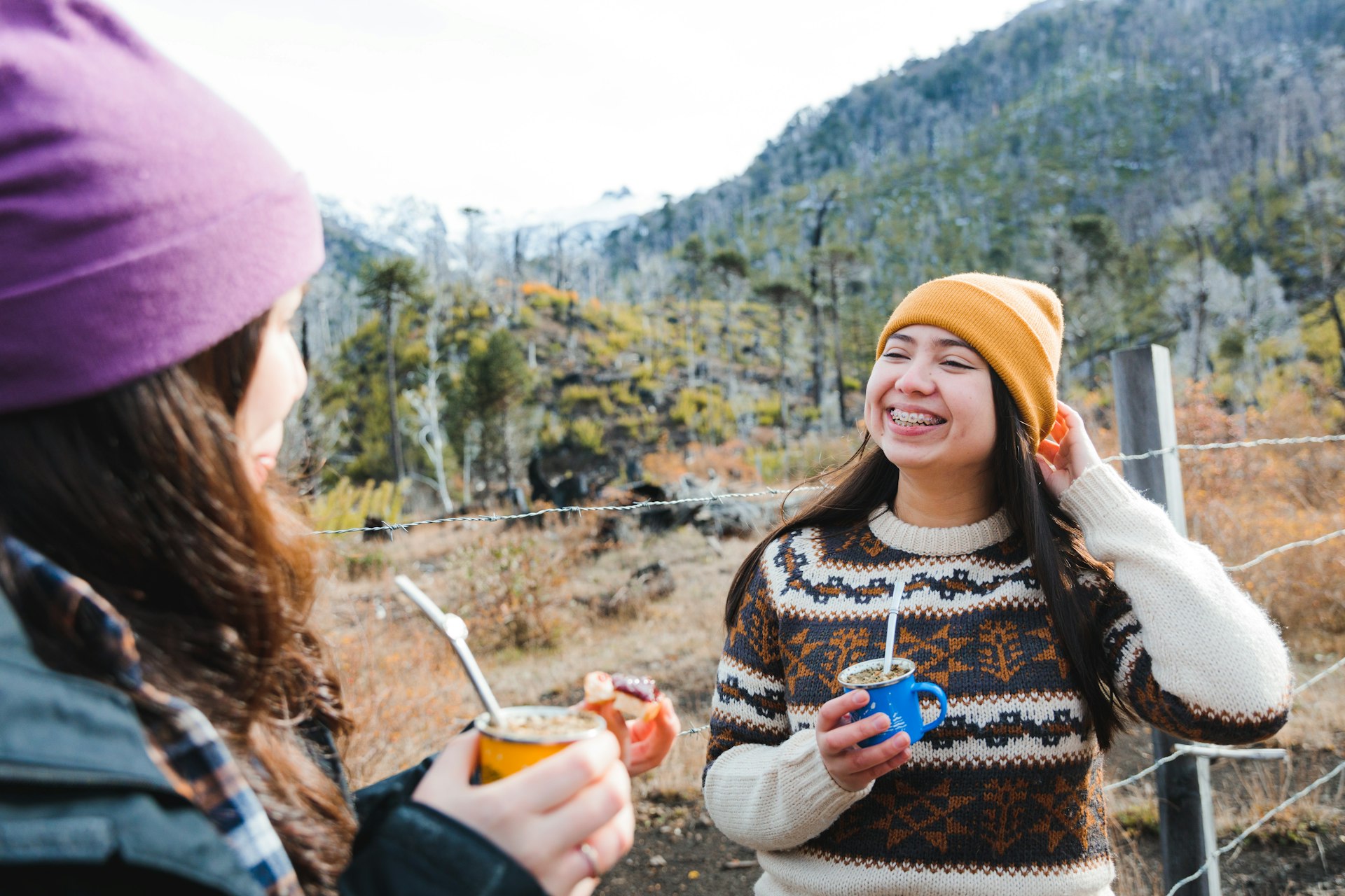 Two young women are smiling and drinking yerba mate outside in the mountainous landscape of Patagonia