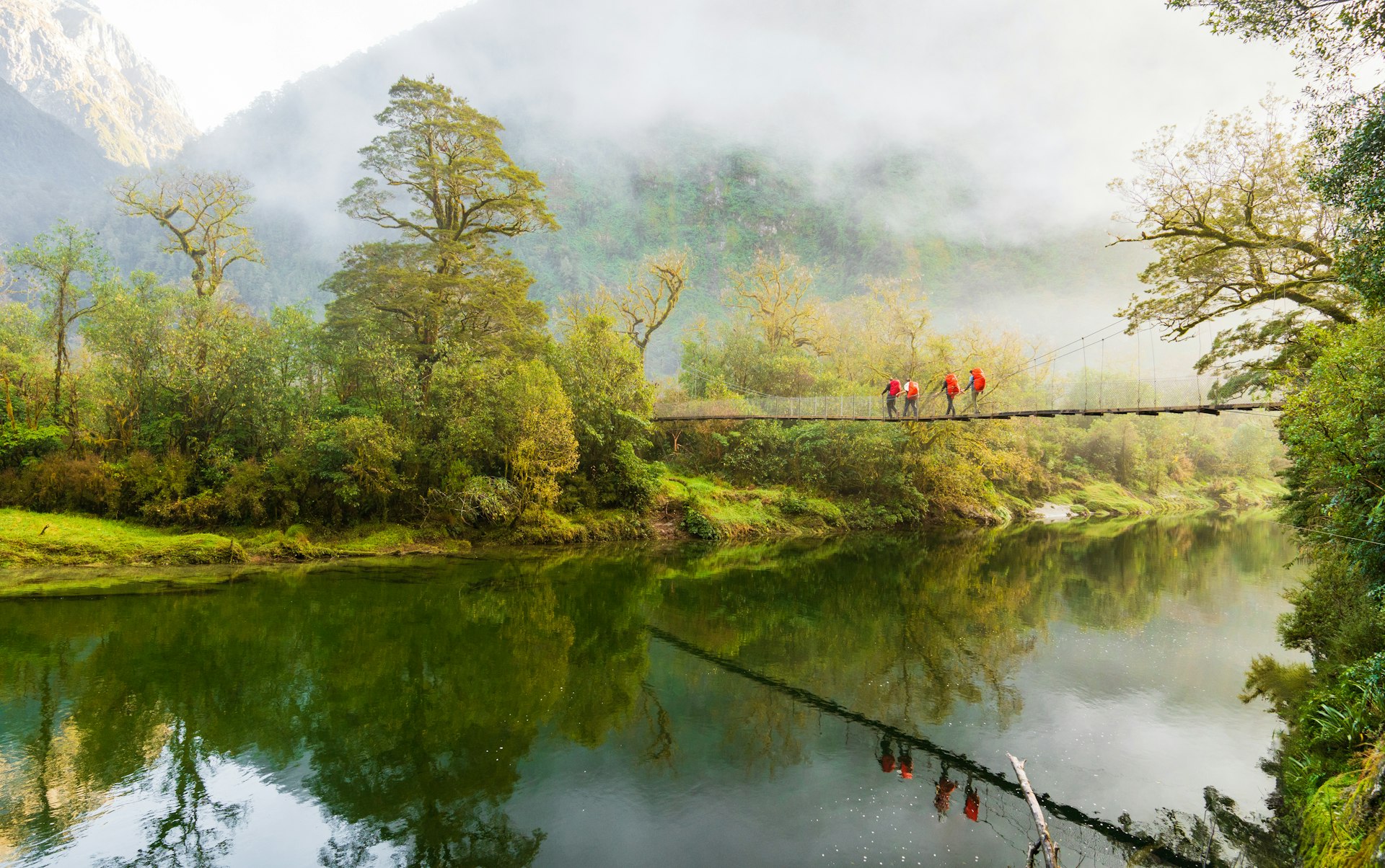 Hikers cross a bridge suspended above a river on a misty day