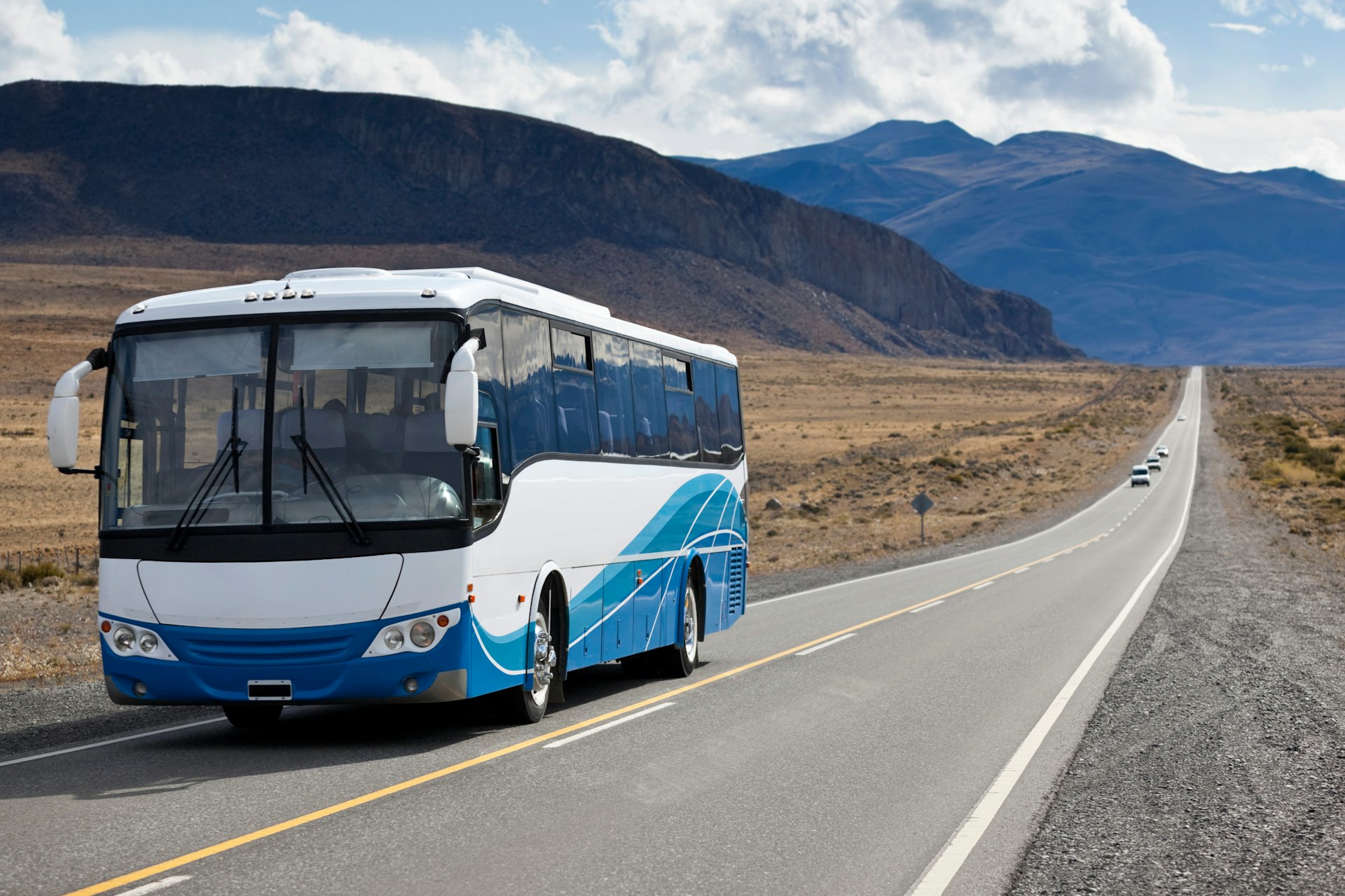 A blue-and-white bus is traveling down a long stretch of highway, with several vehicles in the distance and a backdrop of huge jagged mountains.