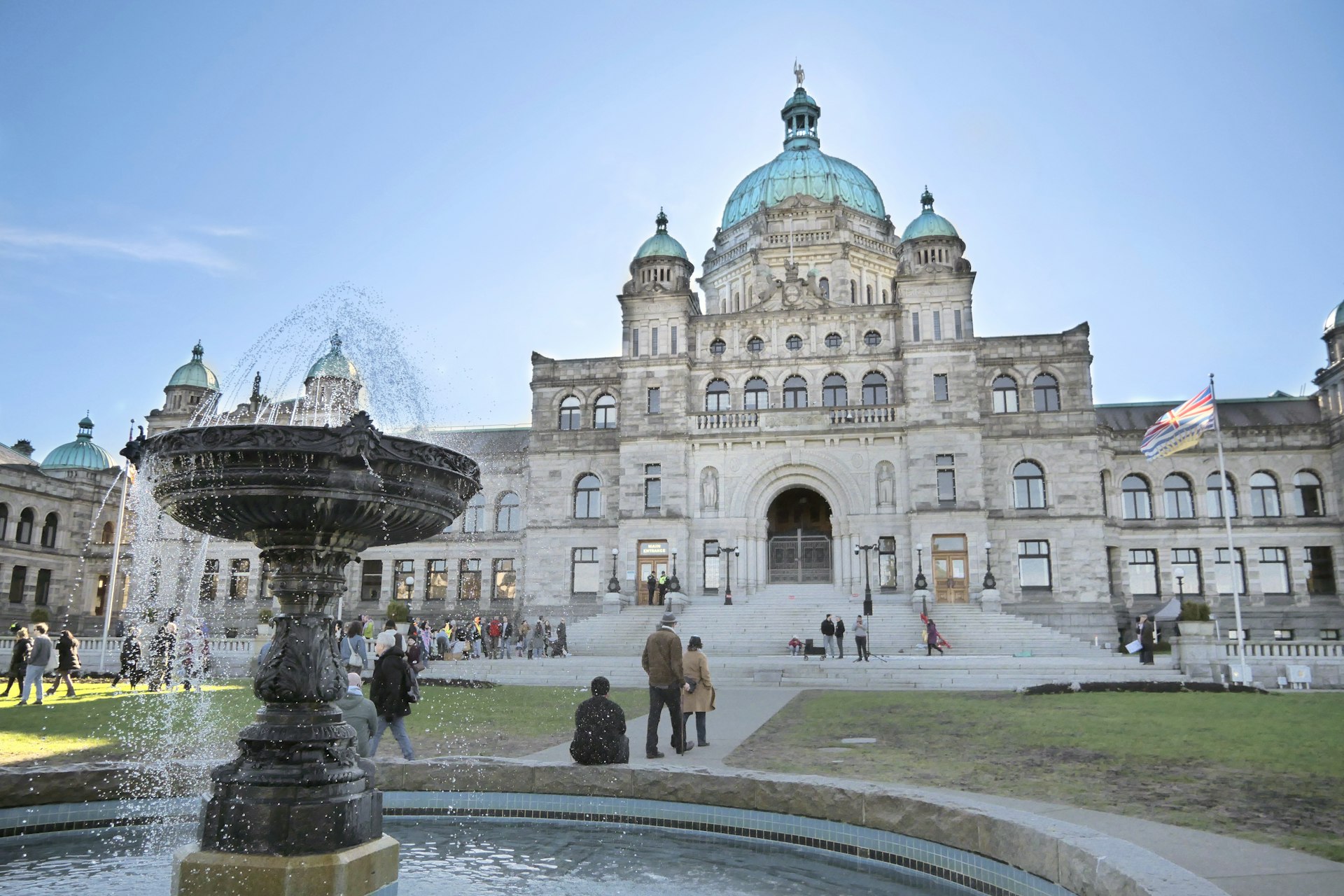 Mant people are milling around beside a fountain outside the Legislative Assembly Parliament Building of British Columbia in Victoria, on a sunny day.