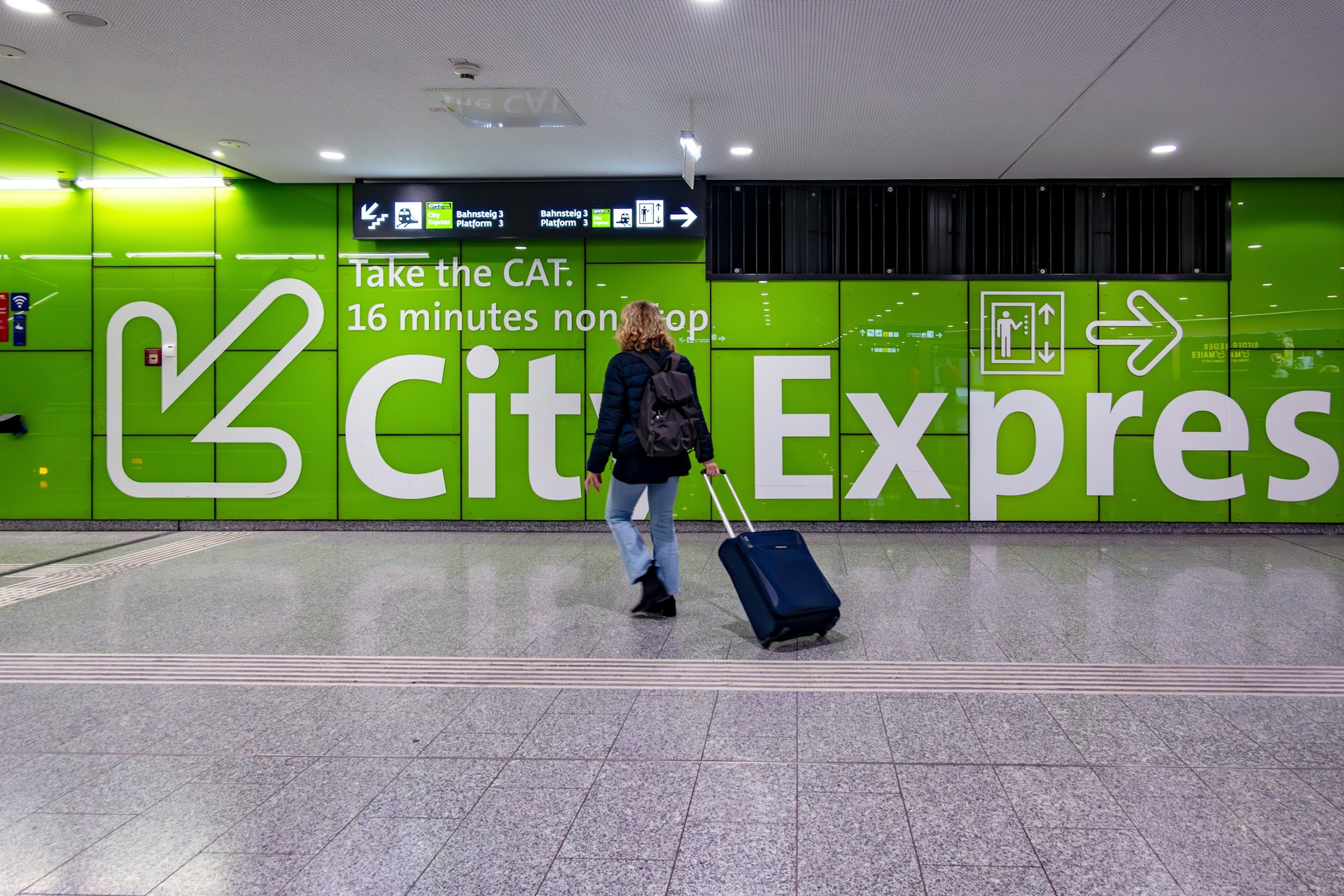 A woman pulling a wheelie suitcase is walking towards a large, lime-green sign pointing towards the City Airport Train, or CAT, in Vienna Airport.
