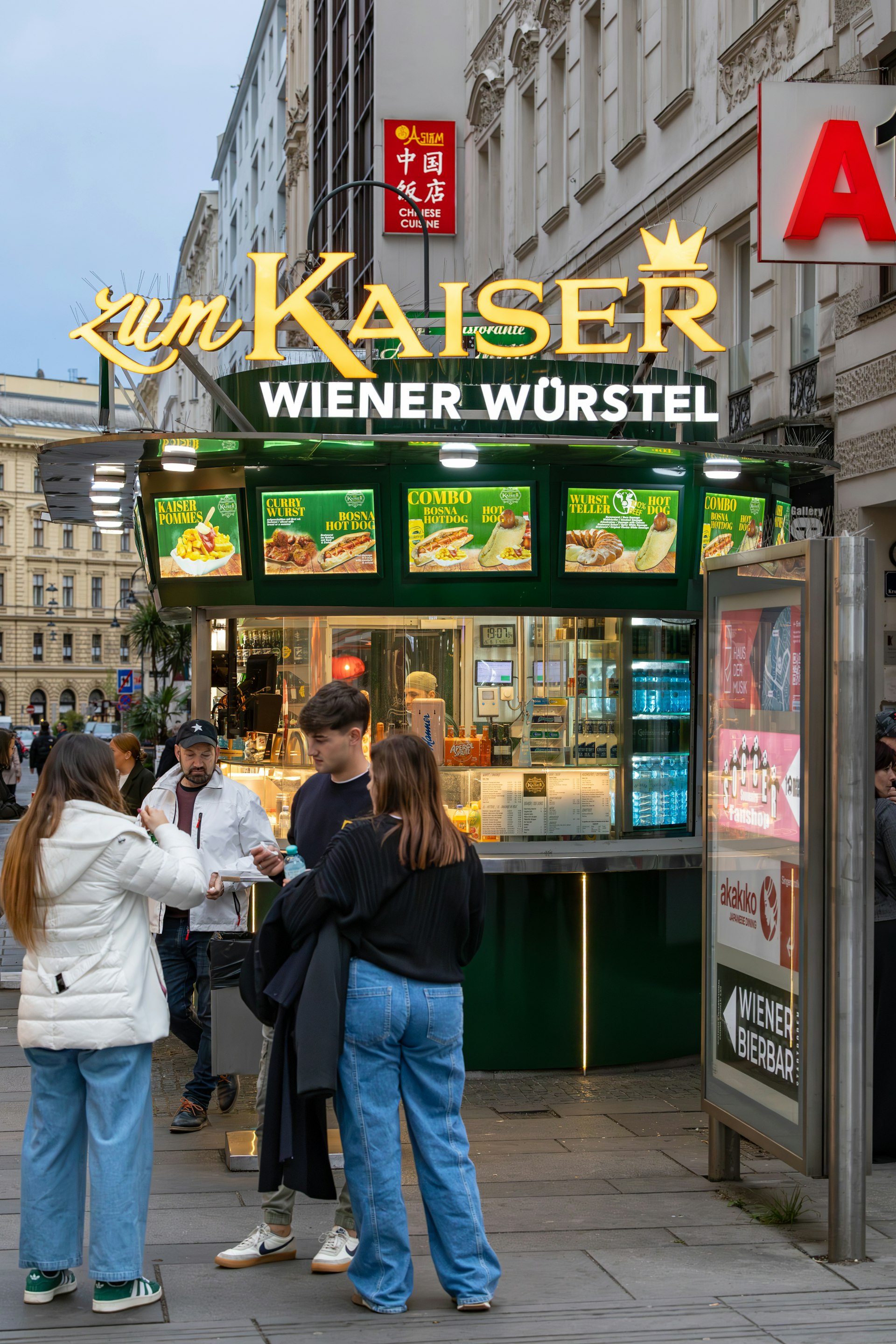 A group of three friends are standing beside a "Würstelstand" (sausage stand) in Vienna