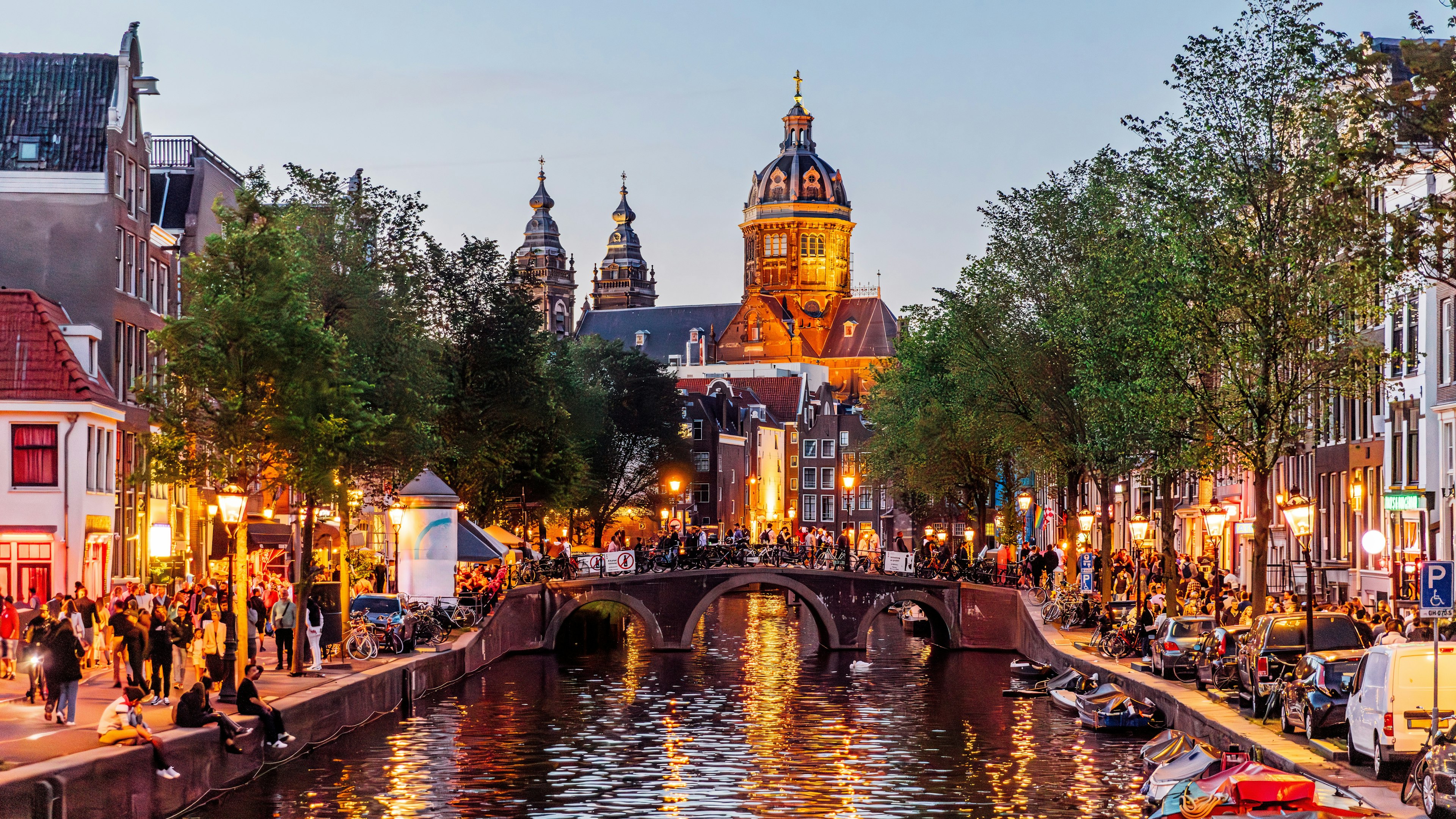 A canal cuts through the centre of Amsterdam's Red Light District, with bars and restaurants (lit in the iconic red lights) flanking the streets either side, while the roads are busy with people.