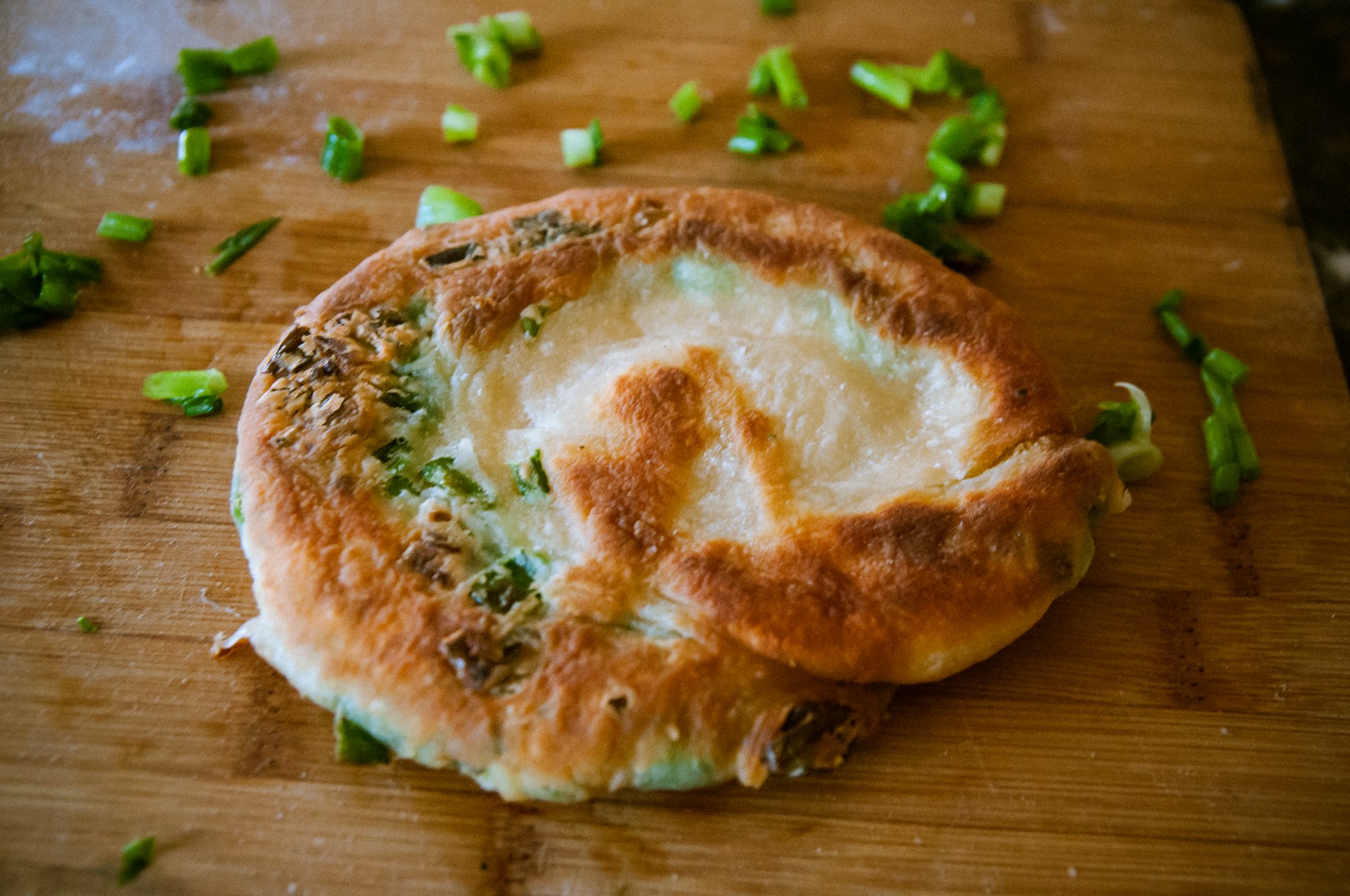 A green onion cake on a wood chopping board with sliced scallions around it.