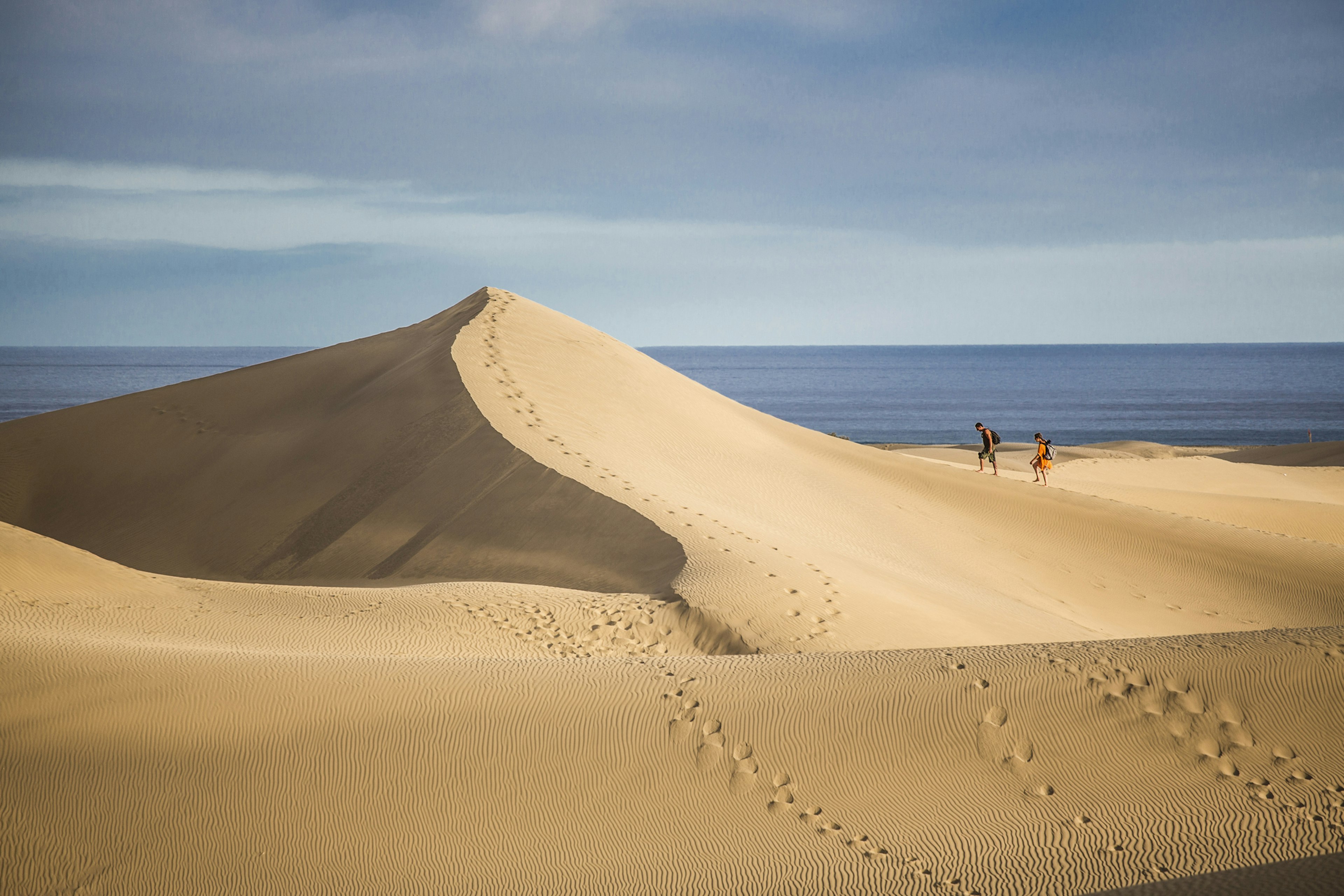 Two people carrying backpacks trudge up the steep slope of a vast sand dune