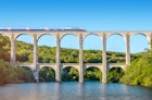 Cize, France - July 9, 2015: French high speed train TGV operated by SNCF, national rail operator on Cize-Bolozon viaduct bridge in Ain, Rhone-Alpes region in France. This train was developed during the 1970s by GEC-Alsthom and SNCF. A TGV test train set the record for the fastest wheeled train, reaching 574.8 km/h (357.2 mph) on 3 April 2007. Viaduct of Cize-Bolozon in summer season in Bugey along Ain river. This viaduct is a combination rail and vehicular viaduct crossing the Ain gorge. An original span built in the same location in 1875 was destroyed in World War II. Reconstructed as an urgent post-war project due to its position on a main line to Paris, the new viaduct reopened in May 1950. It carries road and rail traffic at different levels.
481529752
Brand-name, European Culture, Travel, People Traveling, Tourism, High Speed Train, Locomotive, Elevated Road, High Speed, Arch Bridge, Railway Bridge, Elevated Railway Track, Viaduct, Railroad Crossing, Stone Material, Symmetry, Crossing, Scenics, Arch, Bullet Train, Majestic, Journey, Blue, Ancient, Old, Pattern, French Culture, Architecture, Transportation, Nature, Rural Scene, Panoramic, Ain, Rhone-Alpes, France, Europe, Tree, Summer, Mountain, Hill, Landscape, Sky, River, Water, Railroad Track, Bridge - Man Made Structure, Monument, Train, Mode of Transport, Stone Bridge, SNCF, TGV, Alstom, Bugey