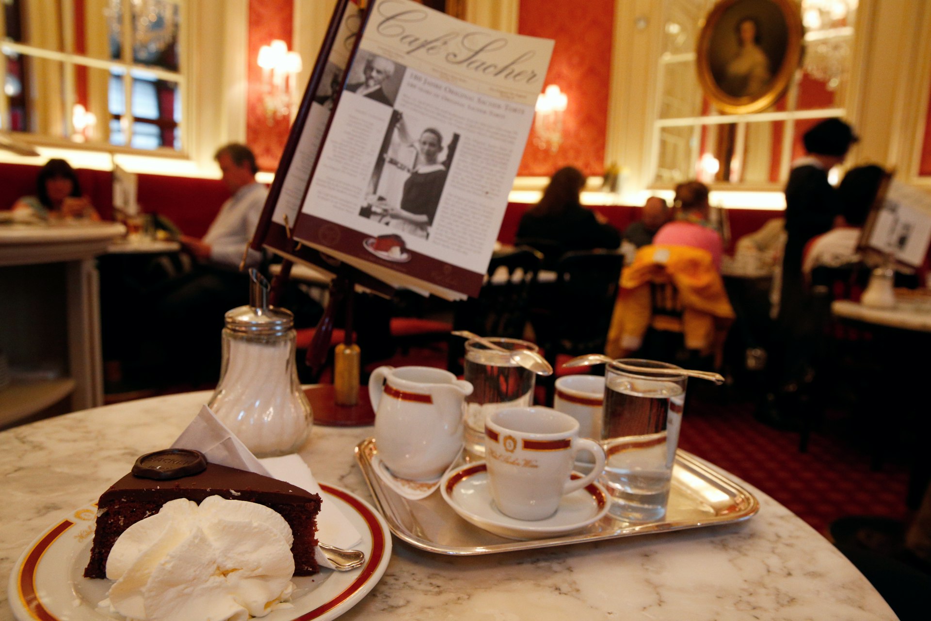 The interior of Café Sacher in Vienna; in the foreground is a table which has a tray holding coffee cups and a milk jug, and next to it is a plate with a slice of Sachertorte