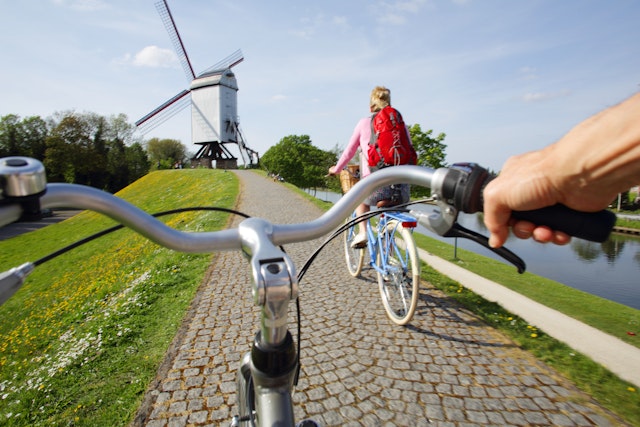 Two people cycling near a windmill, in a shot taken over the handlebars of one rider, Belgium