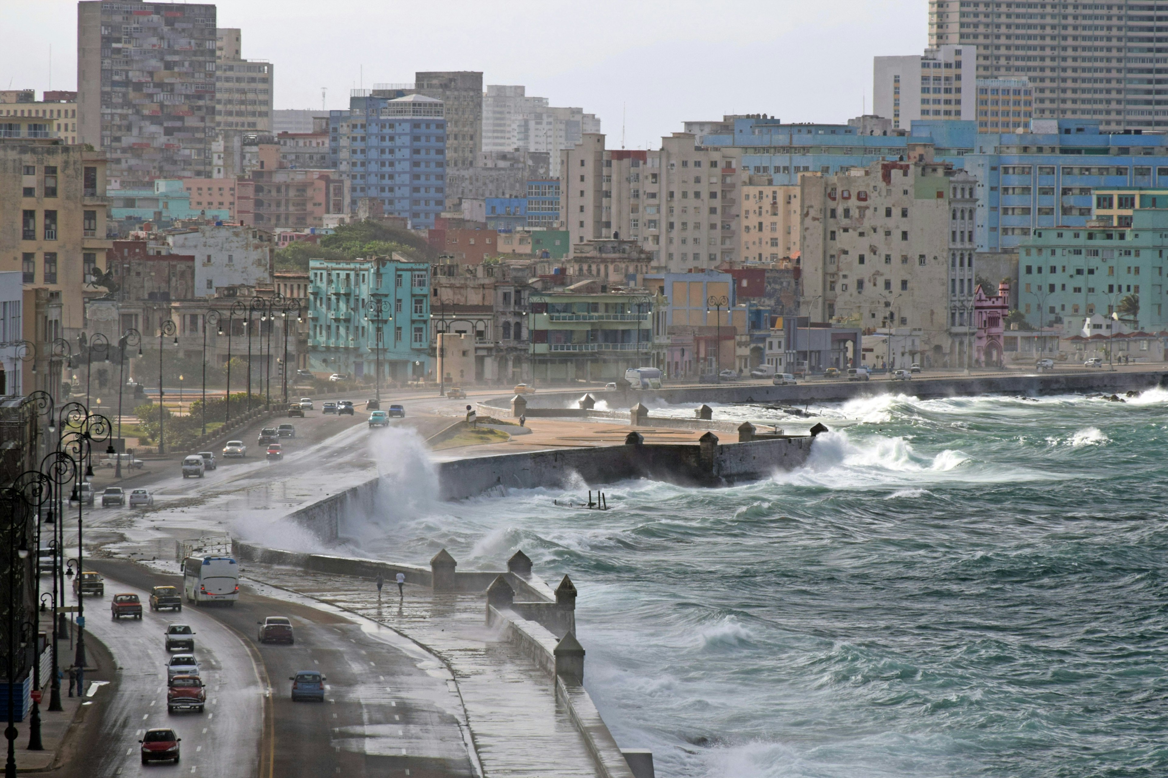 Strong and rough waves hit the shore and wash over the road as cars drive by