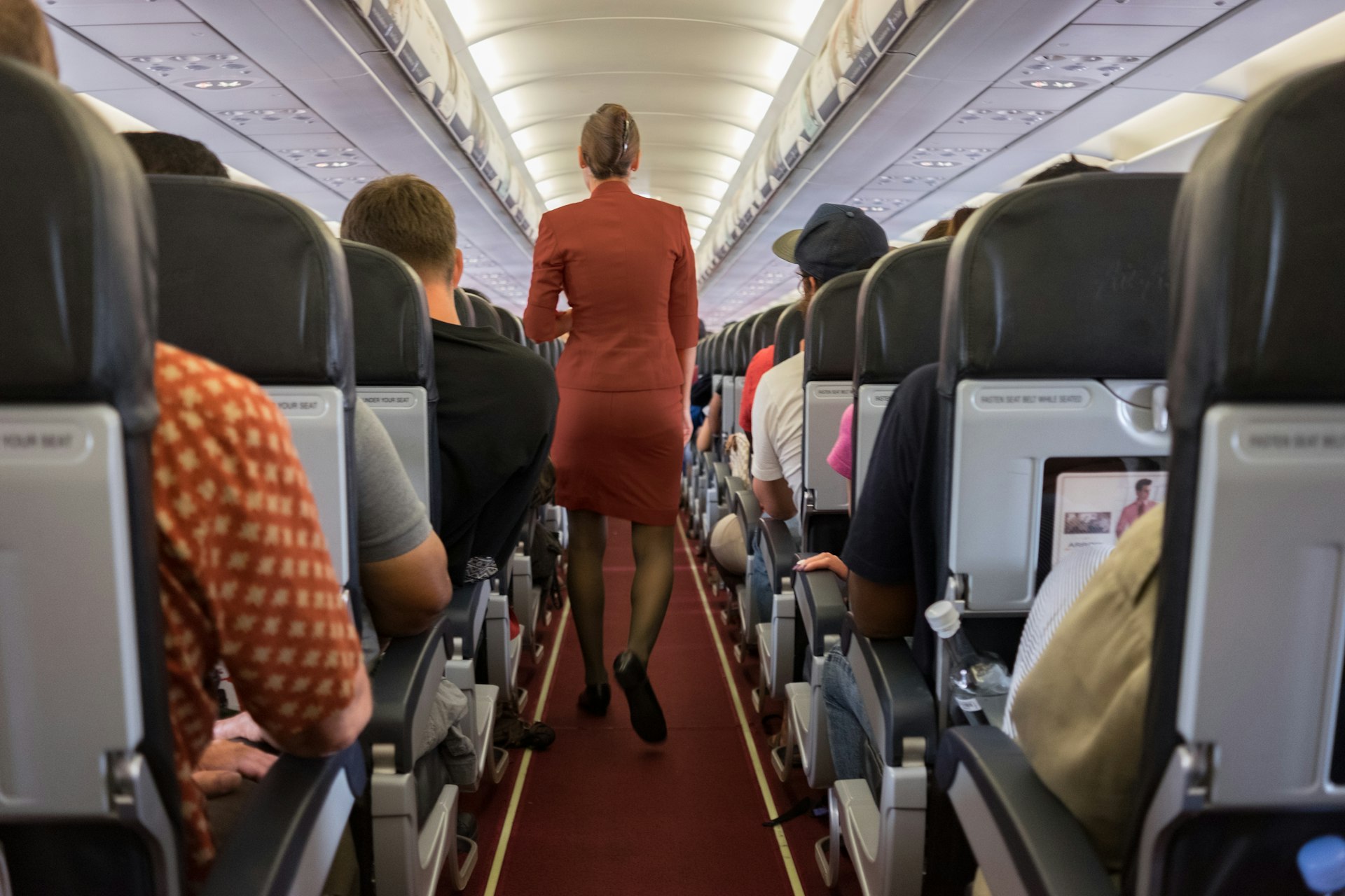 A member of cabin crew wearing a red uniform walks down the central aisle of a plane while all passengers are seated