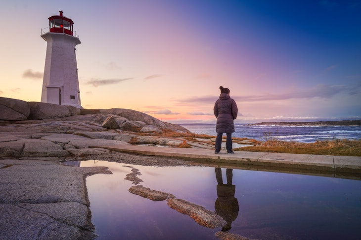 A woman enjoying a winter sunset at Peggys Point Lighthouse, Peggys Cove, Nova Scotia. A reflection is seen in a pool of water gathered on the rocks.