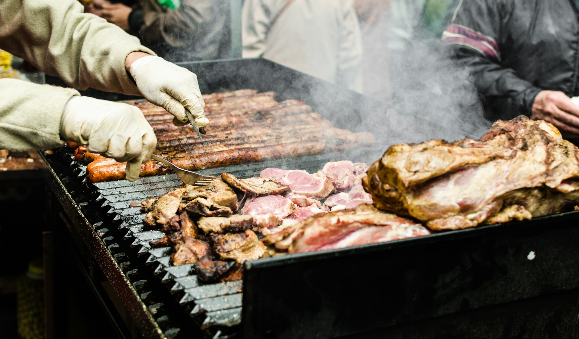 A chef wearing gloves is using tools to turn over a selection of meats on a barbecue grill.