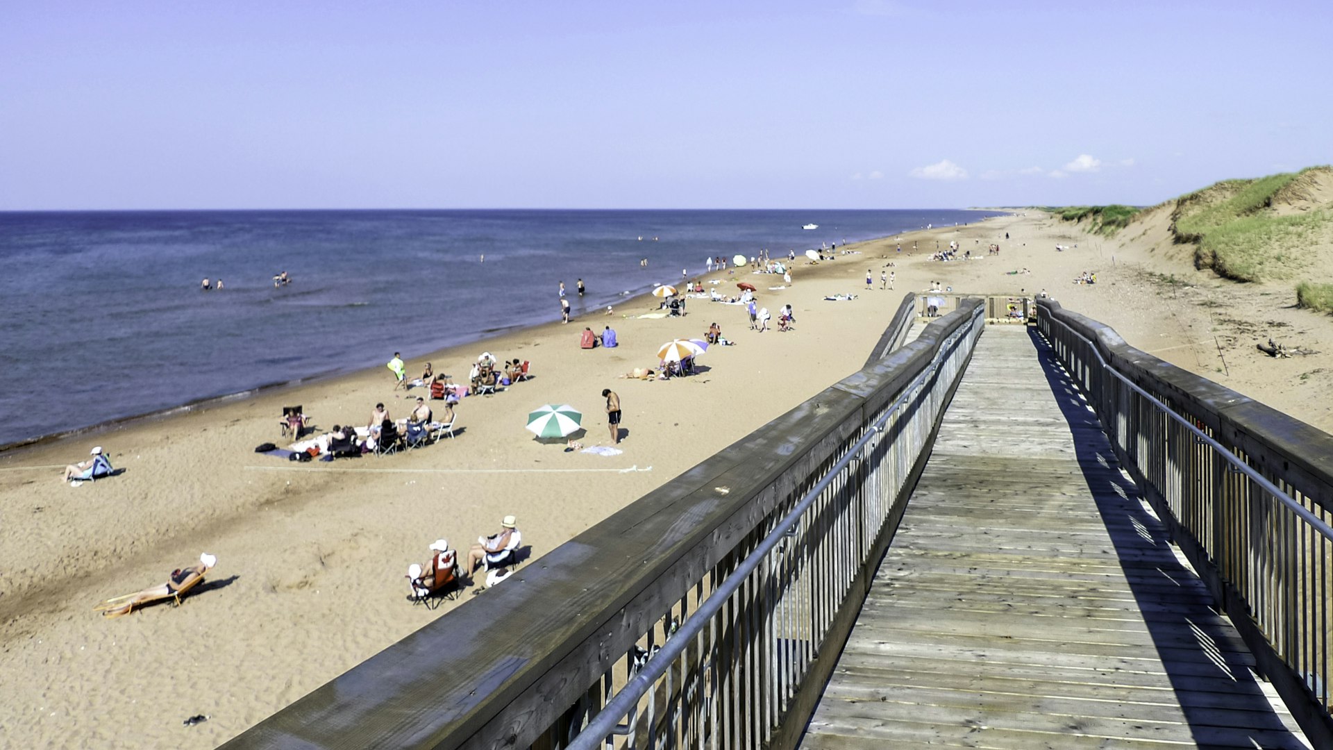 A sloping boardwalk leads down to a sandy beach, where many people are relaxing on the shorefront and swimming in the sea