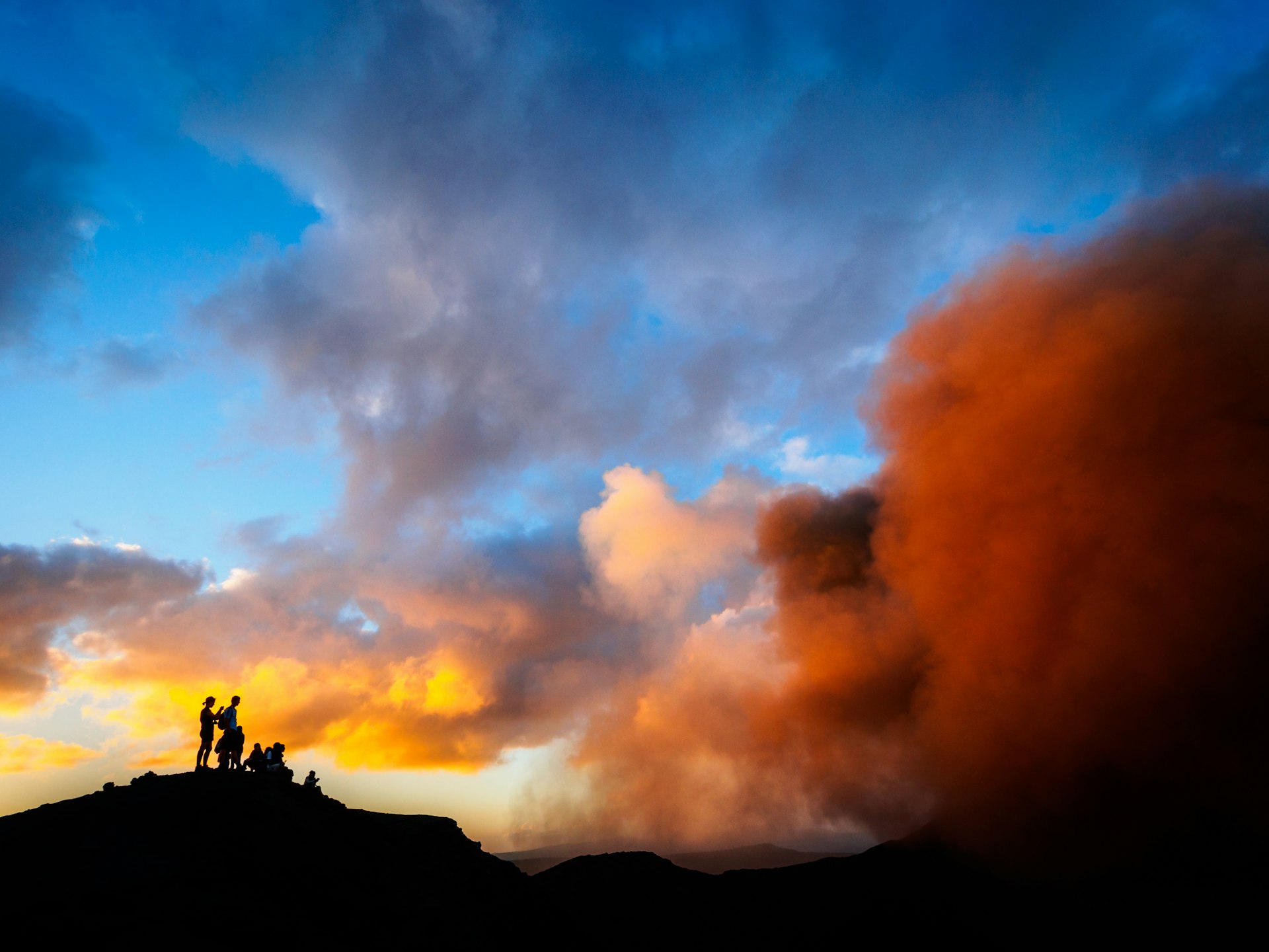 People in silhouette stand at the edge of a volcano crater spewing clouds of dust and smoke against a sunset