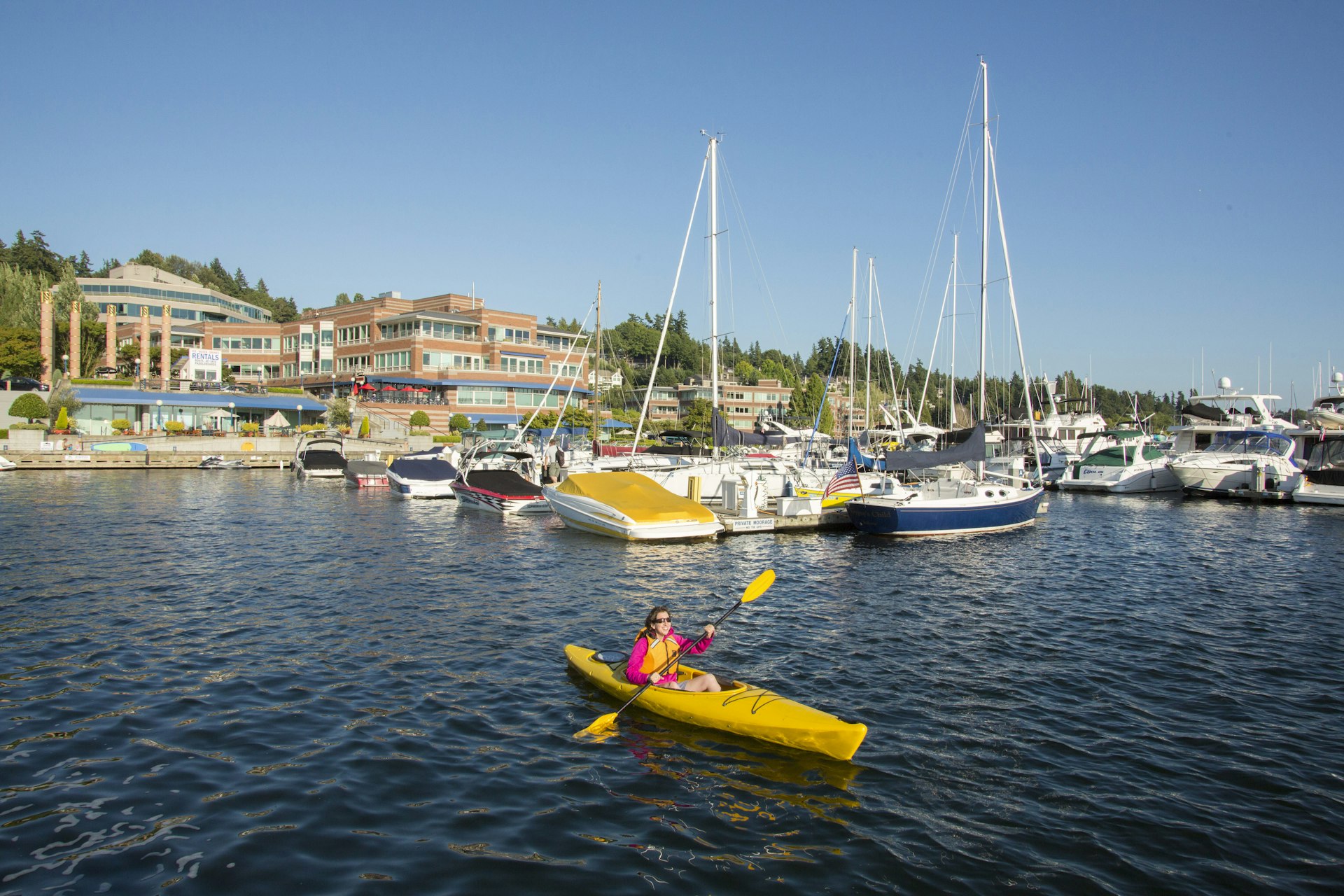 Woman kayaking in a yellow kayak at the Kirkland Marina, Seattle, with buildings in the background. 