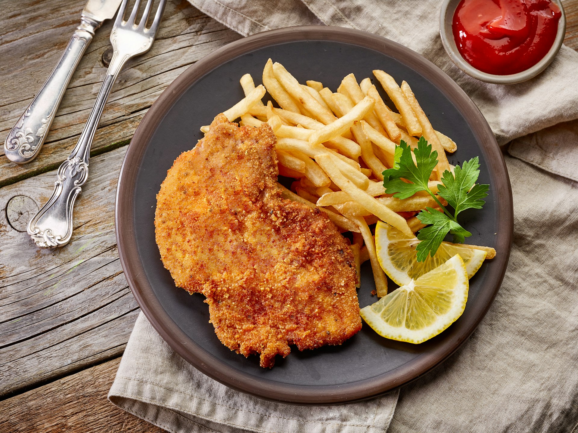 A rustic wooden table with a dinner plate containing schnitzel, French fries and lemon slices