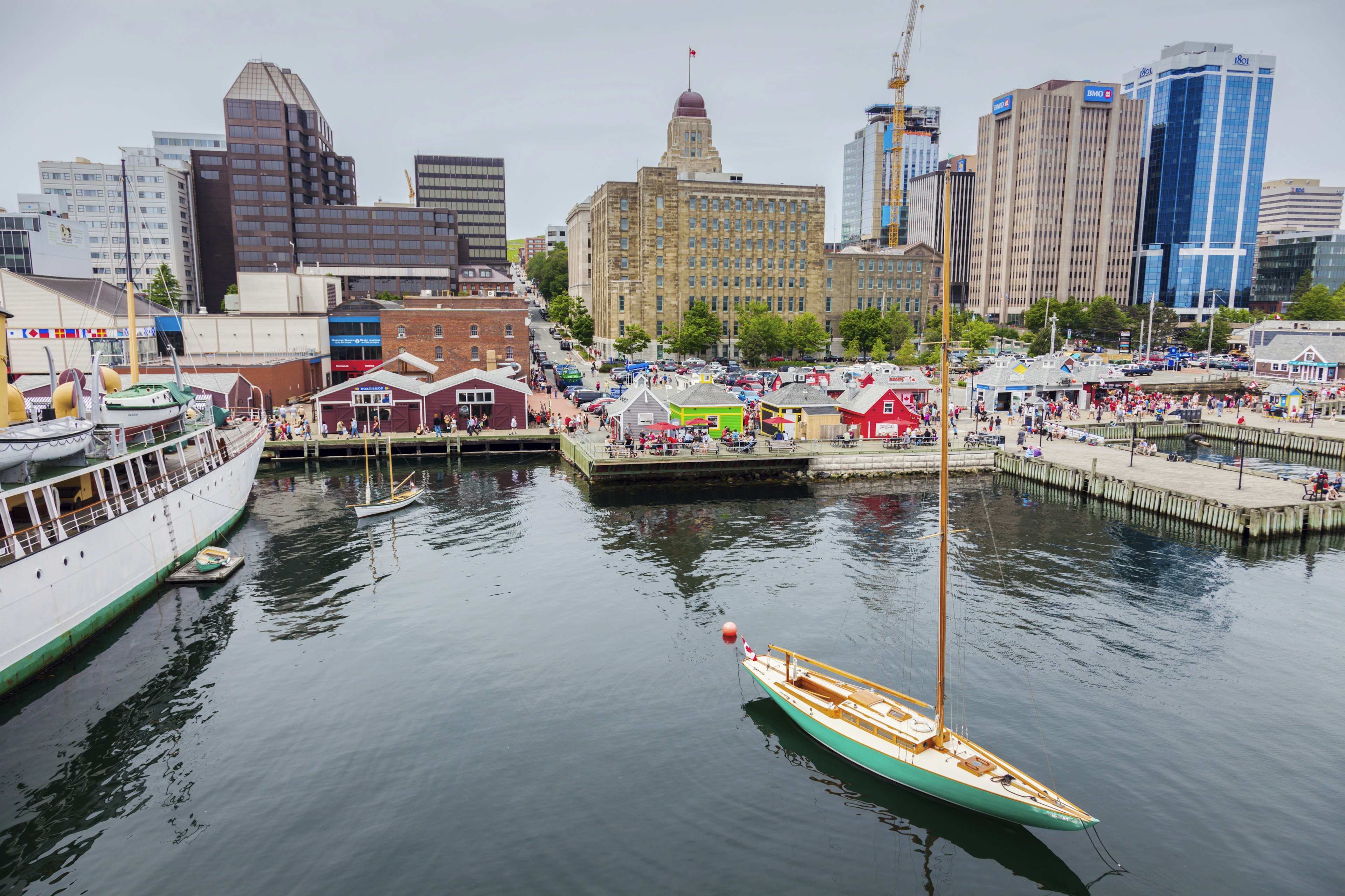 A sail boat leaves a harbor in a city with a busy waterfront