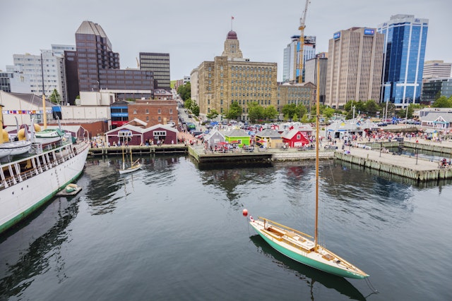 A sail boat leaves a harbor in a city with a busy waterfront