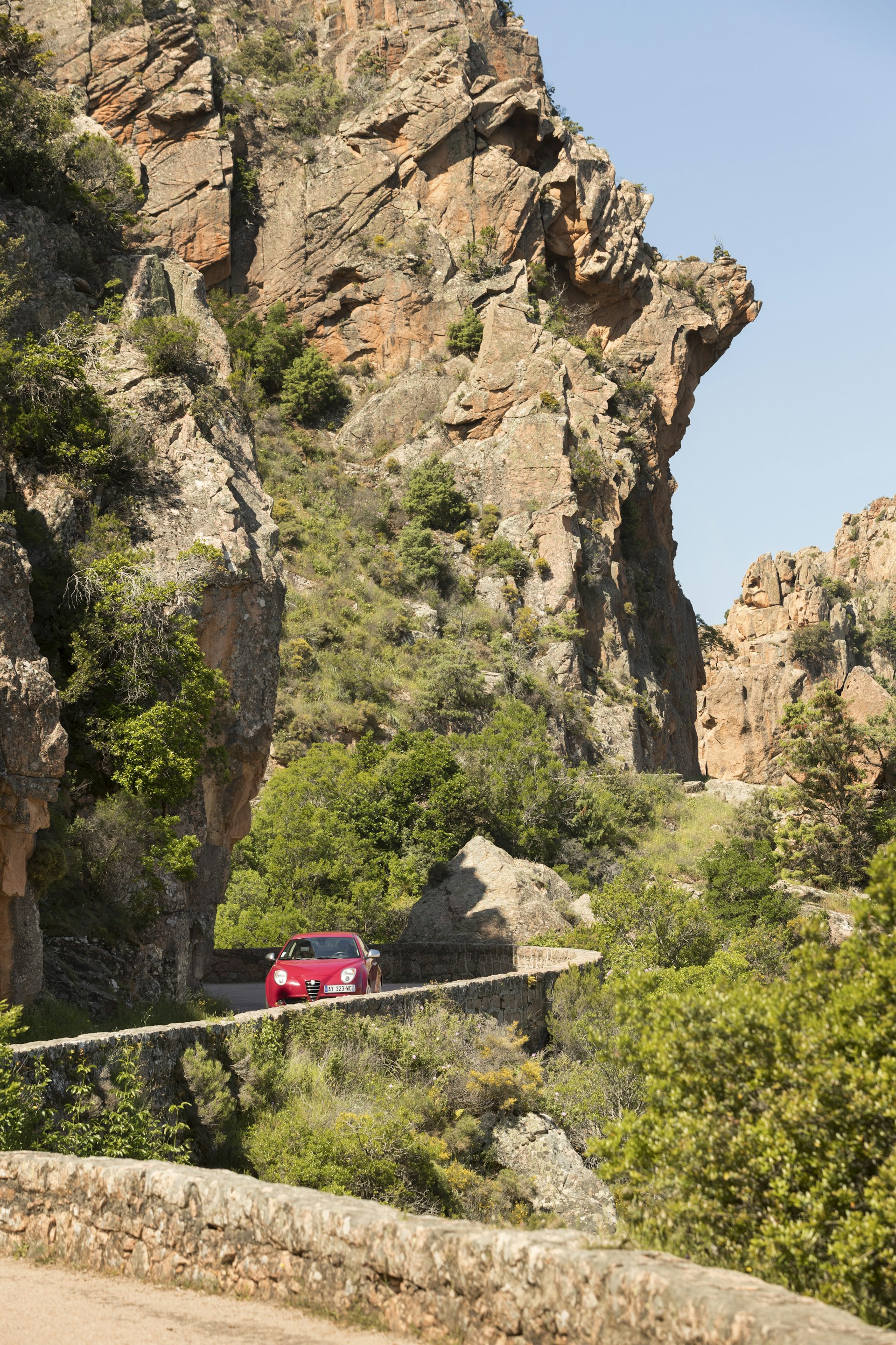 A red car drives around a bend on a narrow road alongside a towering cliff-face