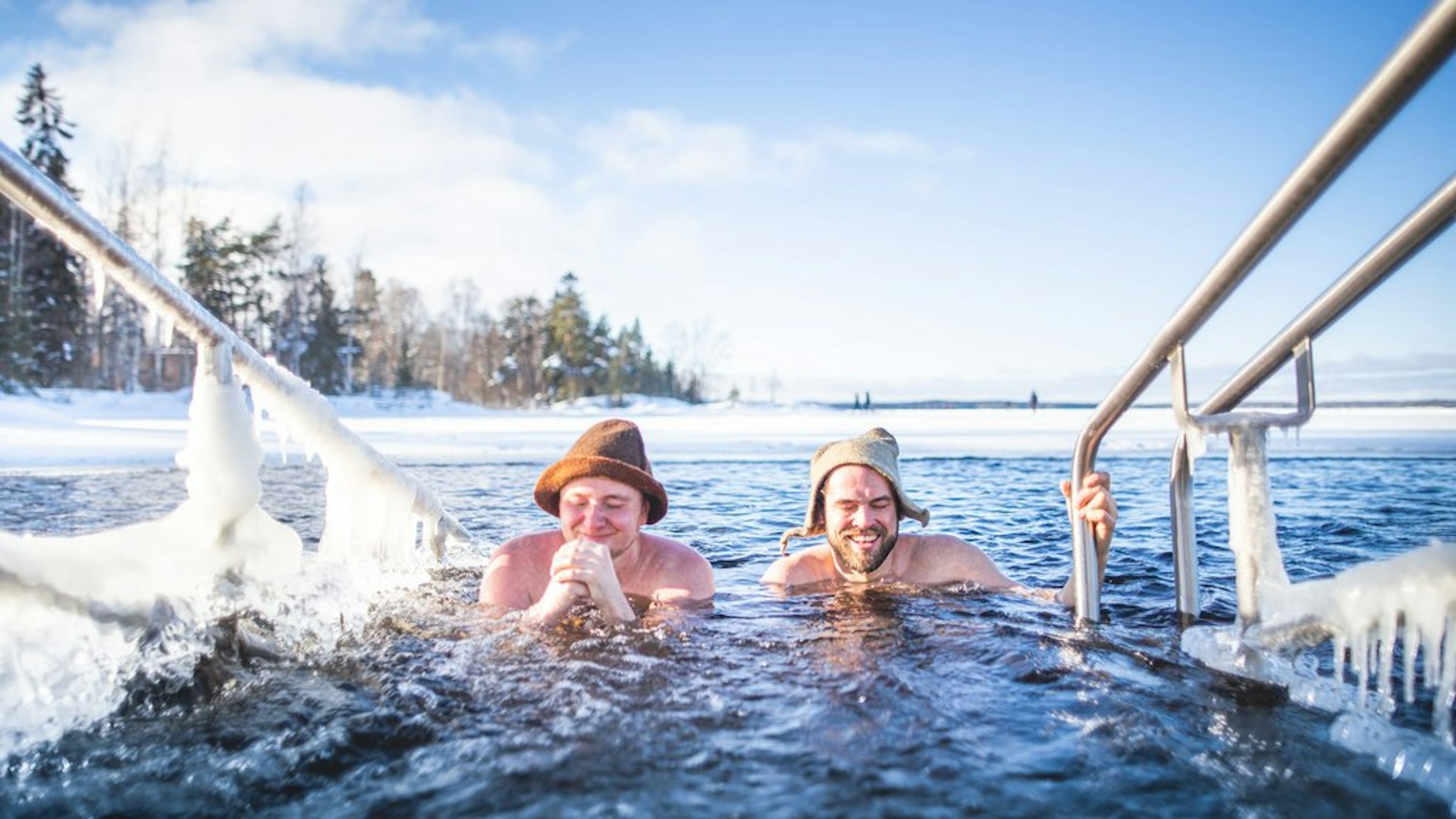 Two men wearing hats are grinning as they submerge their bodies in a frozen pool after having warmed up in a sauna