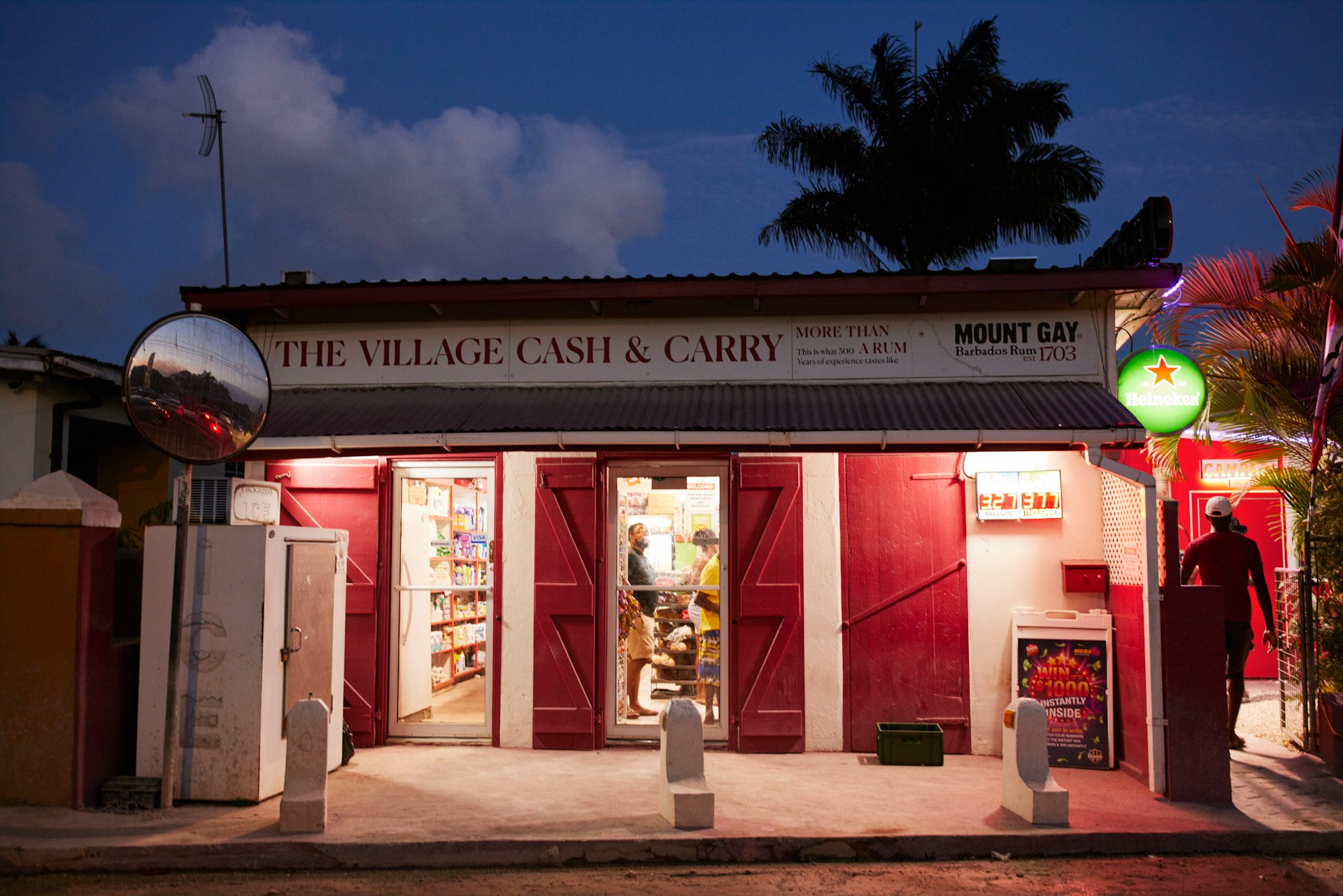 The exterior of a rum shop at night; the sign says "the Village Cash and Carry"