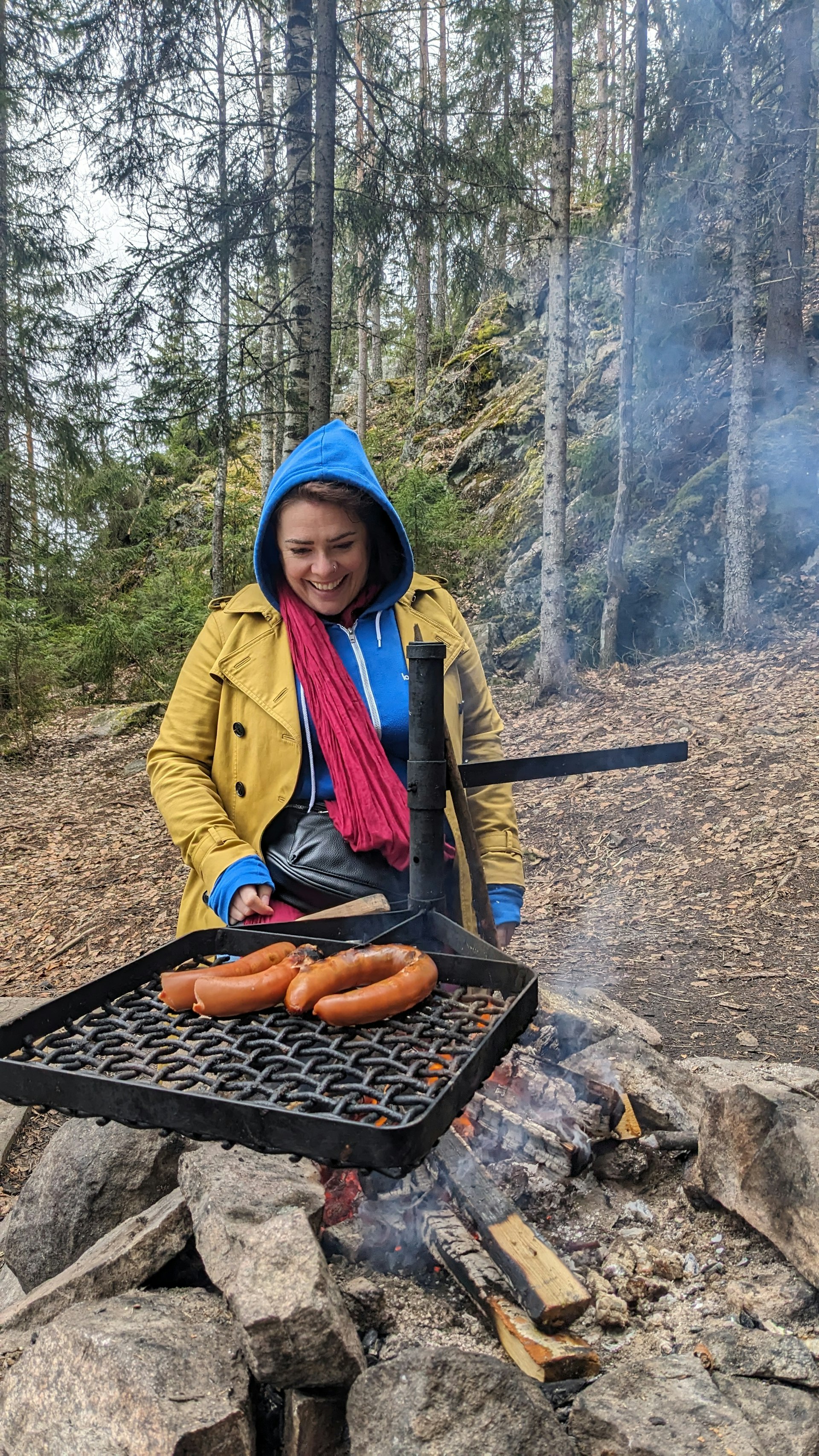 A woman sits near a campfire in woodland watching sausages cook on the grill