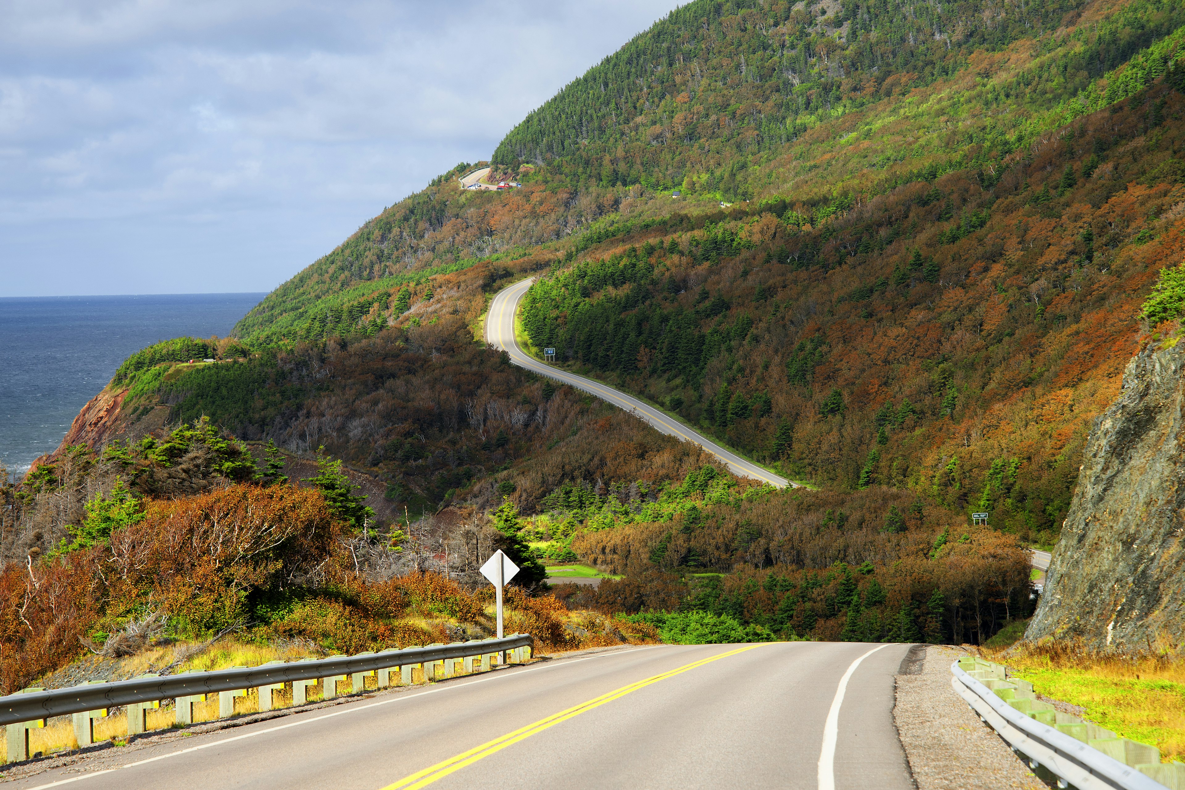 A road unspools with the sea on one side and dramatic fall foliage on the other, Cabot Trail, Cape Breton, Nova Scotia, Canada