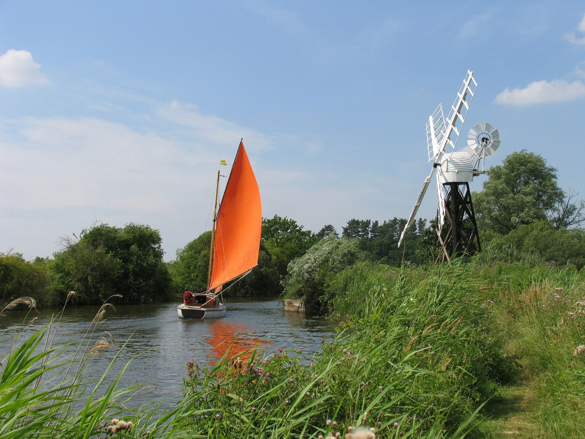 A sailboat passes a windmill near The How Hill Trust and River Ant on the Norfolk Broads, Ludlum, Norfolk.