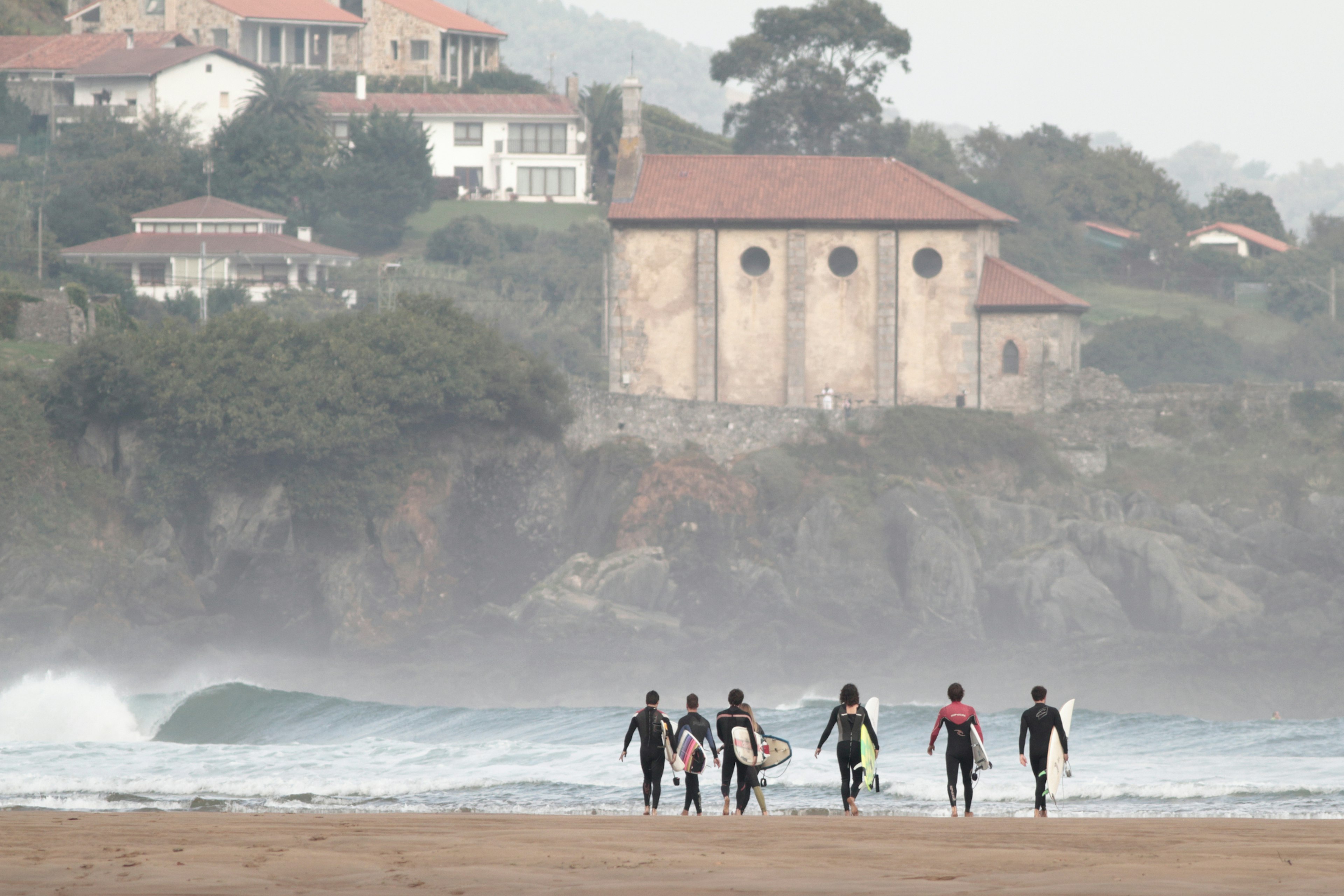 A group of surfers carrying boards stride towards the surf. A large church and houses line the distant hillside