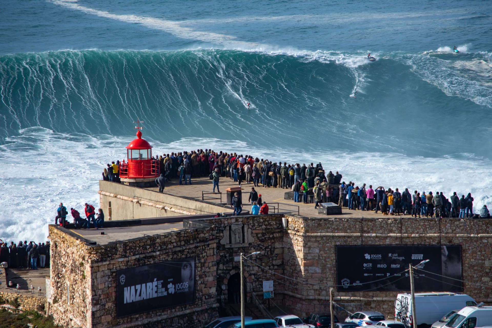 Pessoas se alinham em uma passarela que leva a um farol para que possam observar as enormes ondas que estão sendo surfadas perto da costa