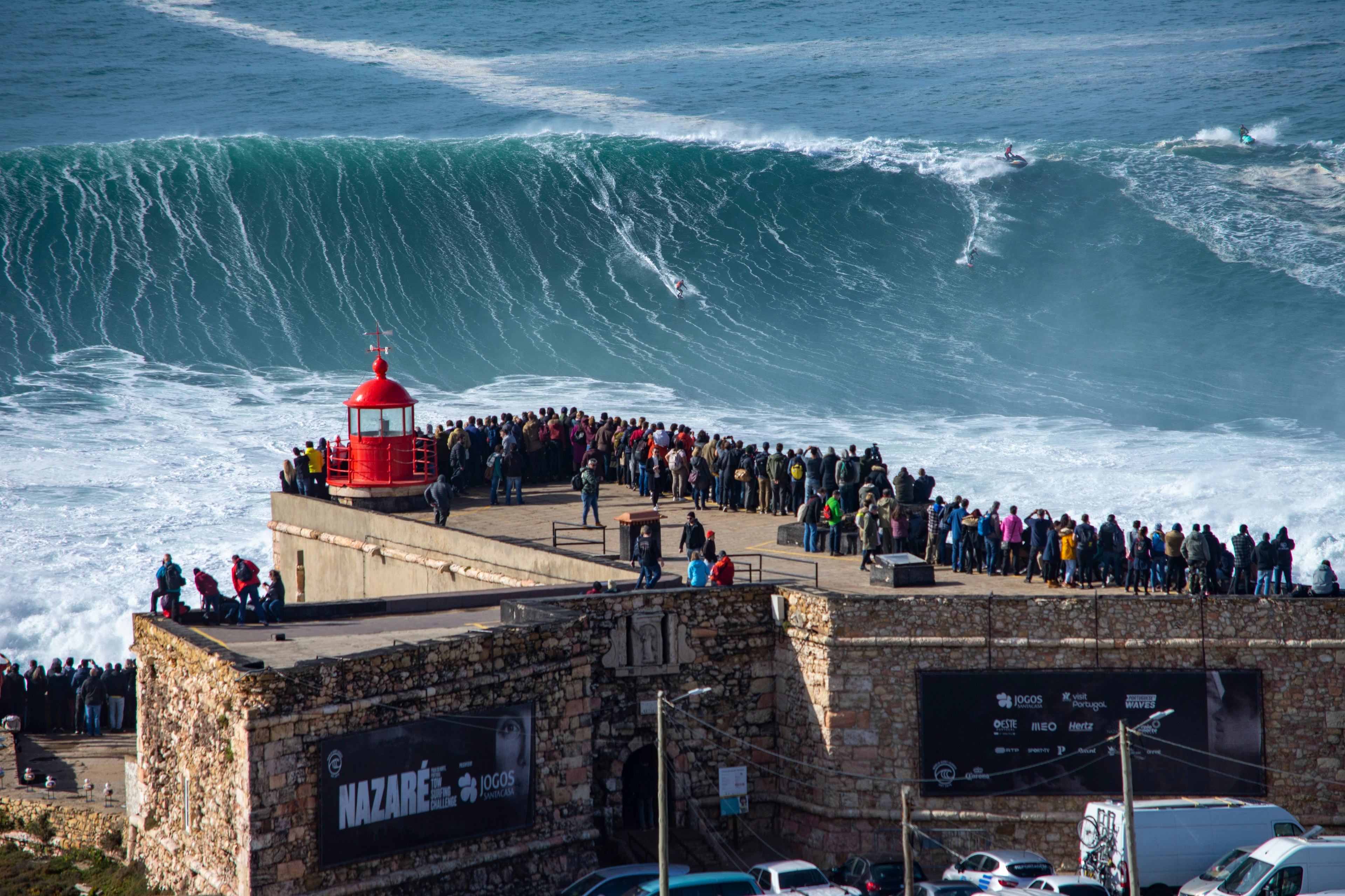 People line a walkway leading to a lighthouse so they can watch the huge waves that are being surfed near the shore