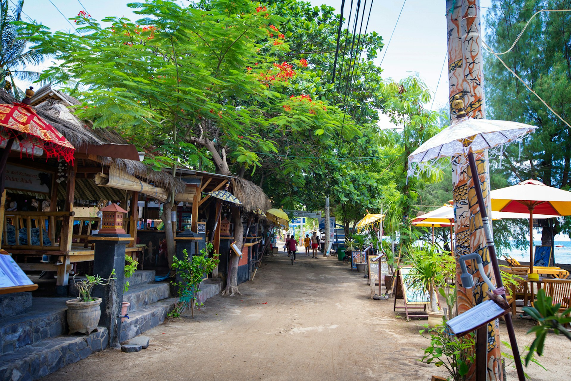 People wander down a shop- and restaurant-lined pathway near a beach