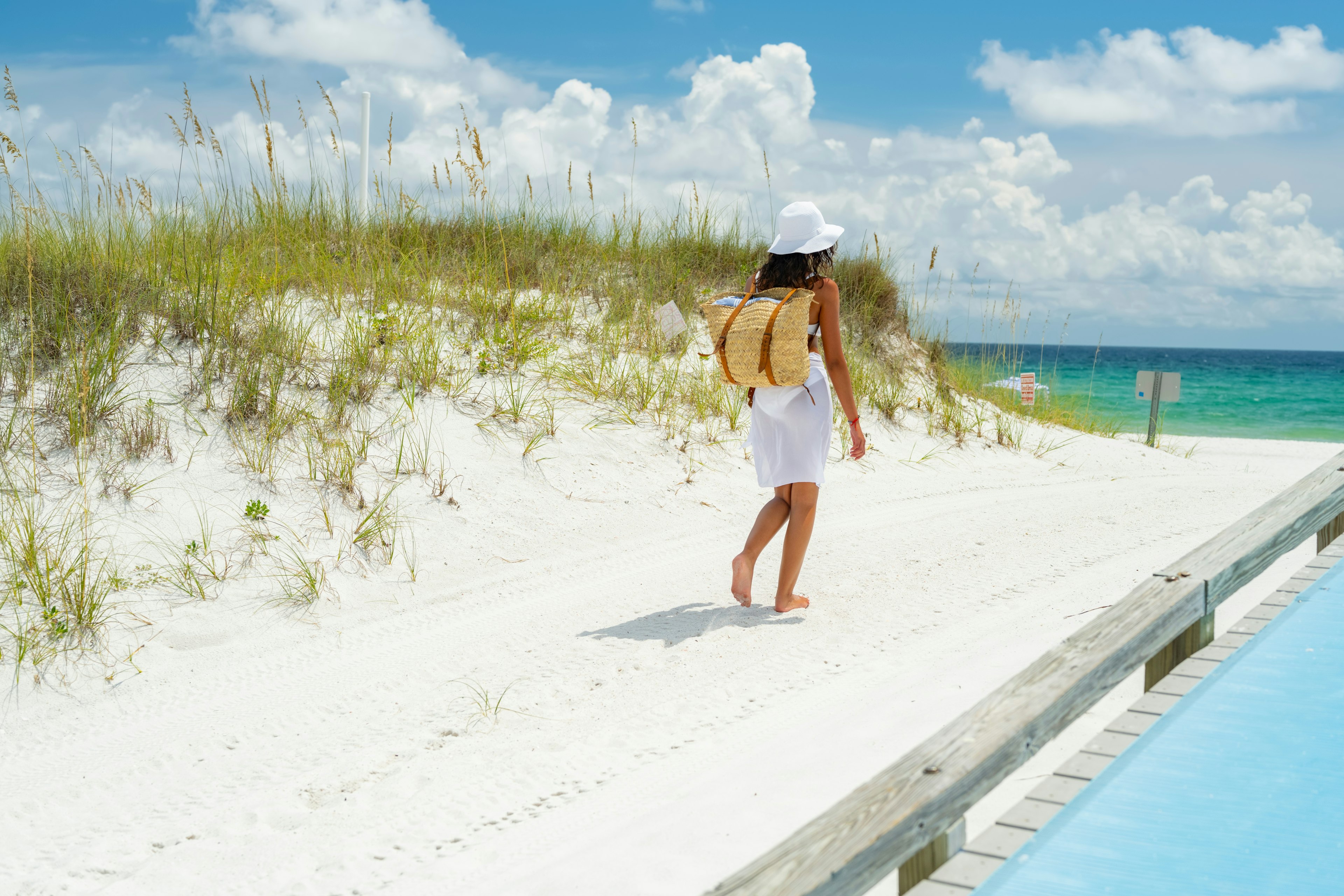 A woman in a white bikini on white sandy beach emerald waters of the emerald coast the Gulf Of Mexico with a hat and a drink and beach bag ;