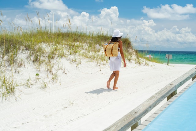 A woman in a white bikini on white sandy beach emerald waters of the emerald coast the Gulf Of Mexico with a hat and a drink and beach bag ;