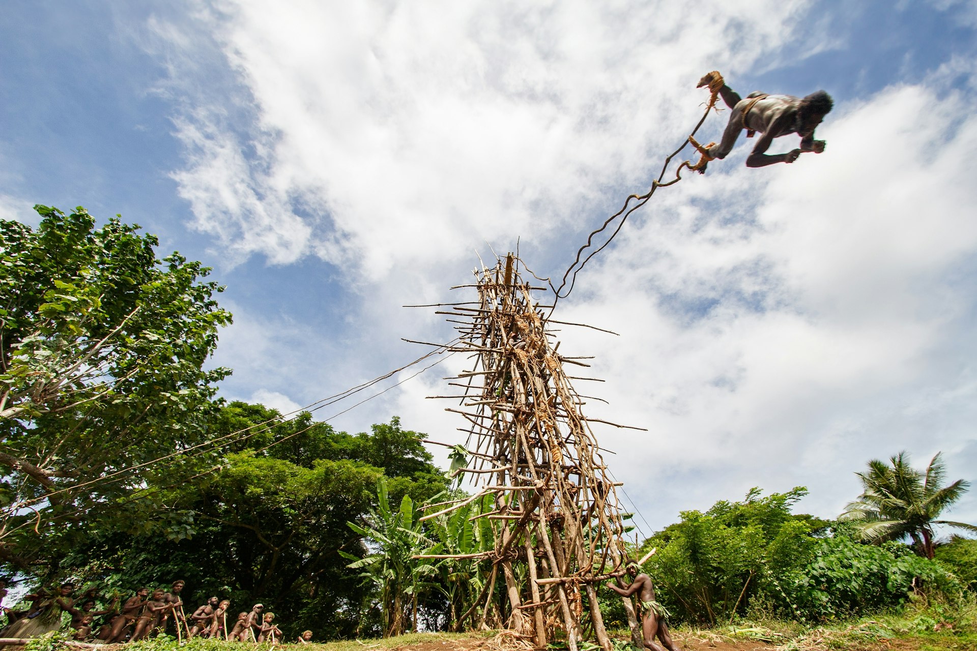A person with vines attached to their leg flies down towards the ground from a makeshift tower