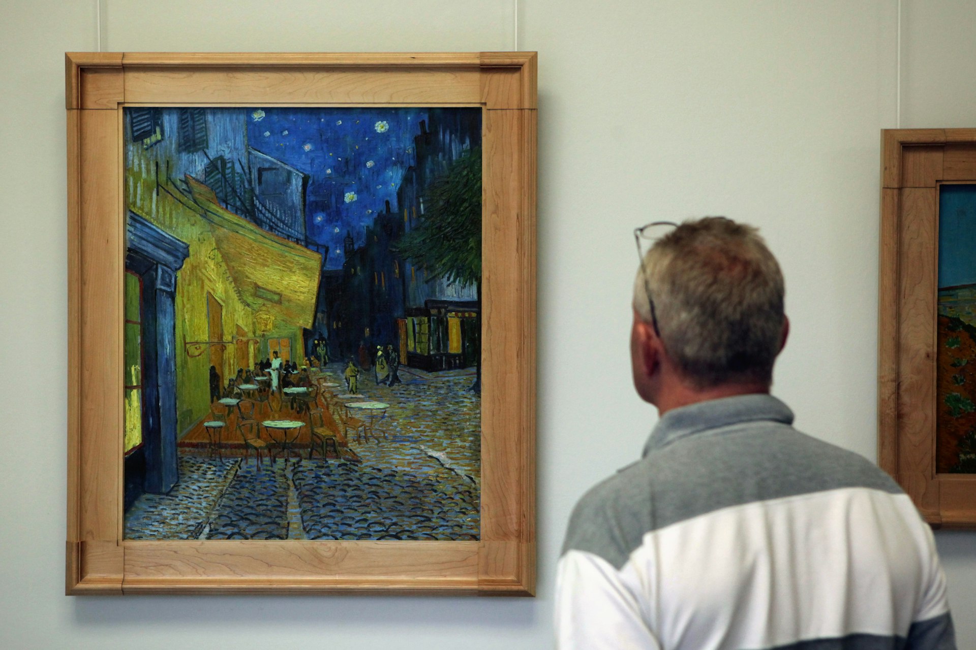 A man stands and looks at a framed painting hanging on a wall of people outside a cafe at night