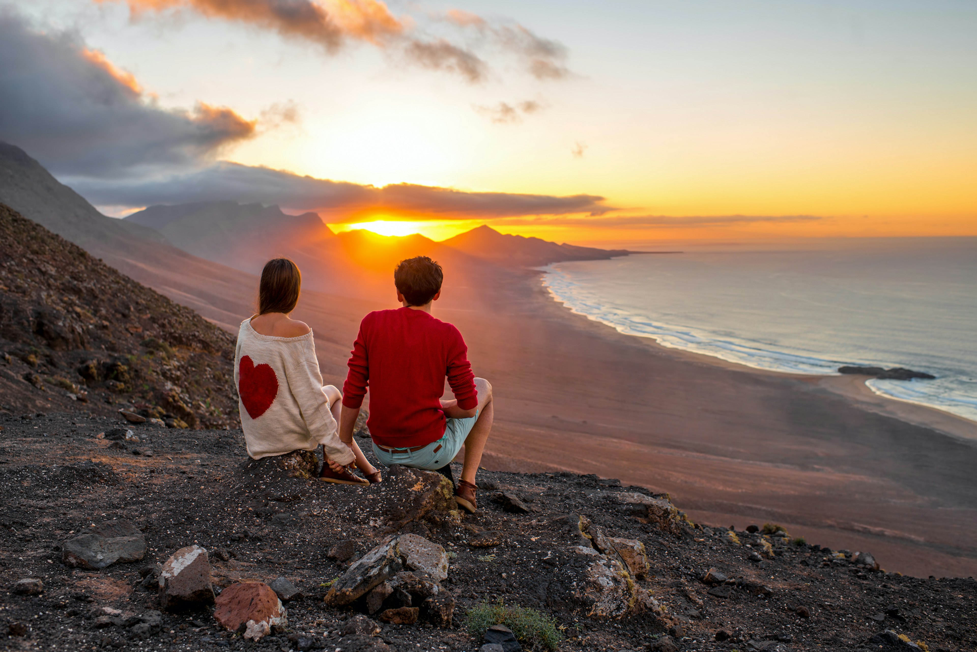 A couple sit together on a volcanic outcrop above a beach watching the sunset.