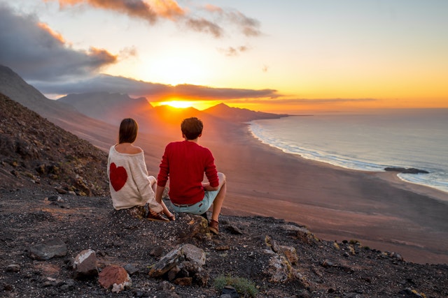 Young couple enjoying beautiful sunset sitting together on the mountain with great view on Cofete coastline on Fuerteventura island.