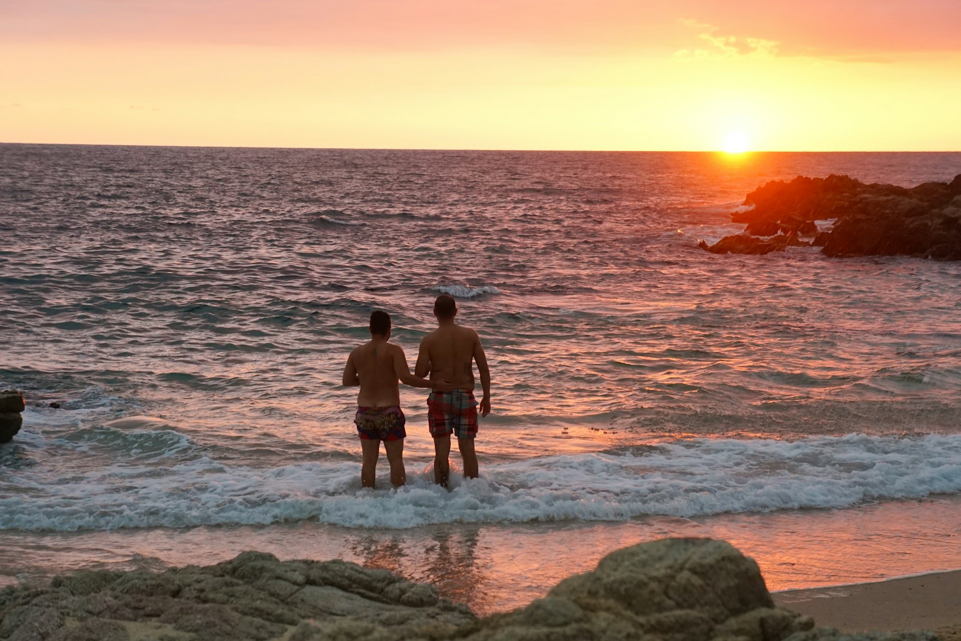 Two men stand in the surf at sunset at Conchas Chinas beach, Puerto Vallarta, Jalisco, Mexico